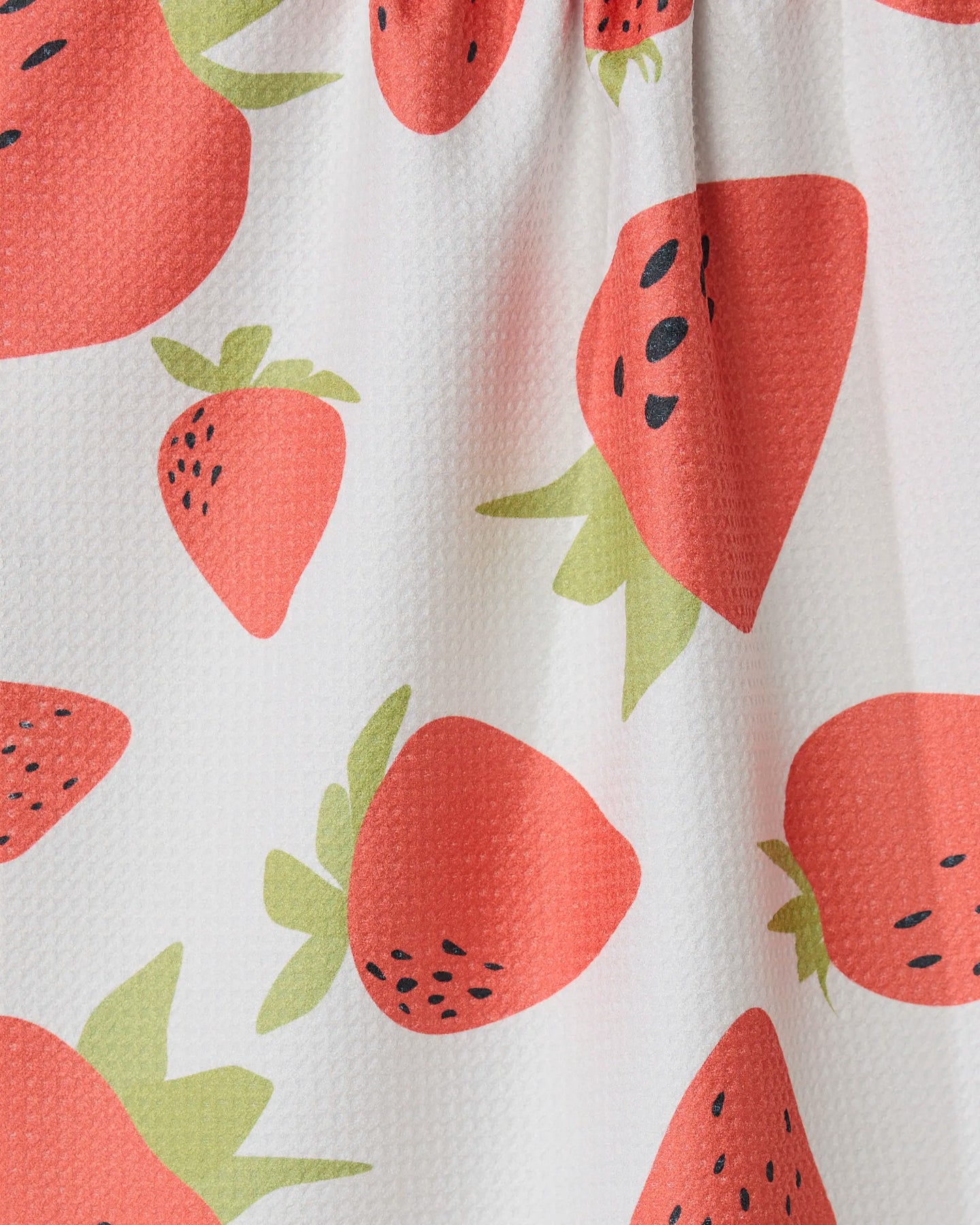 Close-up of GEOMETRYs Sunny Strawberries kitchen towel, made from white recycled fabric and featuring a red strawberry pattern with green leaves and black seeds. The ultra-absorbent towel has a textured look and a gathered seam at the top.