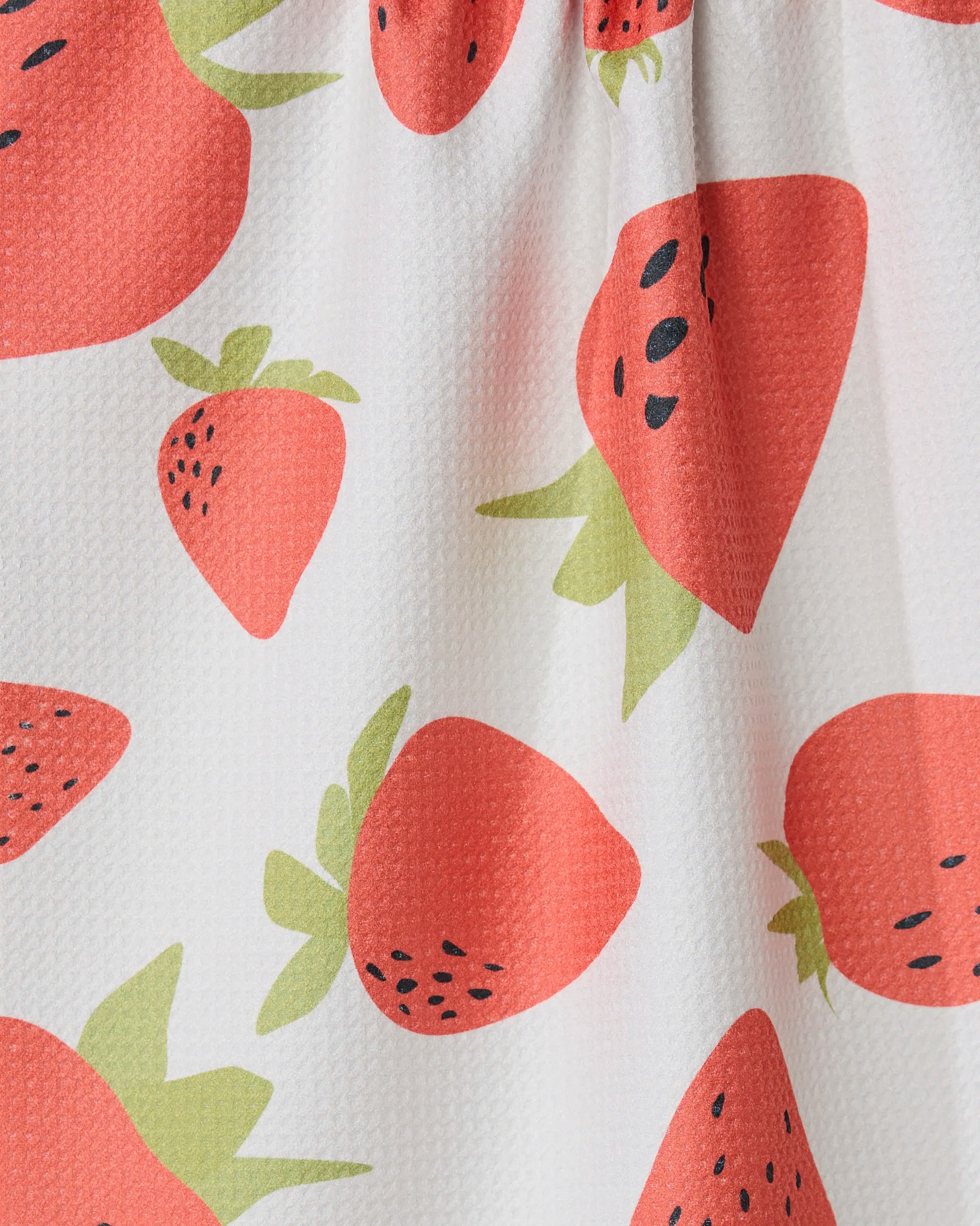 Close-up of GEOMETRYs Sunny Strawberries kitchen towel, made from white recycled fabric and featuring a red strawberry pattern with green leaves and black seeds. The ultra-absorbent towel has a textured look and a gathered seam at the top.
