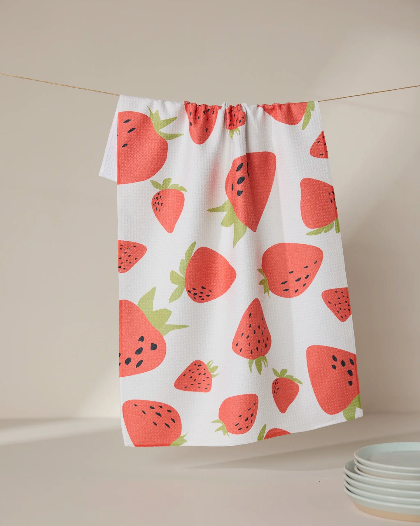 The GEOMETRY Sunny Strawberries tea towel, featuring a colorful strawberry pattern on ultra-absorbent white fabric, hangs on a string against a neutral background with white plates in the lower right corner.