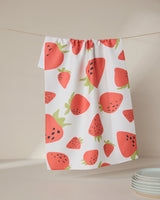 The GEOMETRY Sunny Strawberries tea towel, featuring a colorful strawberry pattern on ultra-absorbent white fabric, hangs on a string against a neutral background with white plates in the lower right corner.