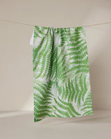 The Swaying Leaf tea towel by GEOMETRY, featuring a green and white repeating fern motif and made from recycled materials, is displayed hanging on a thin string against a neutral beige background.