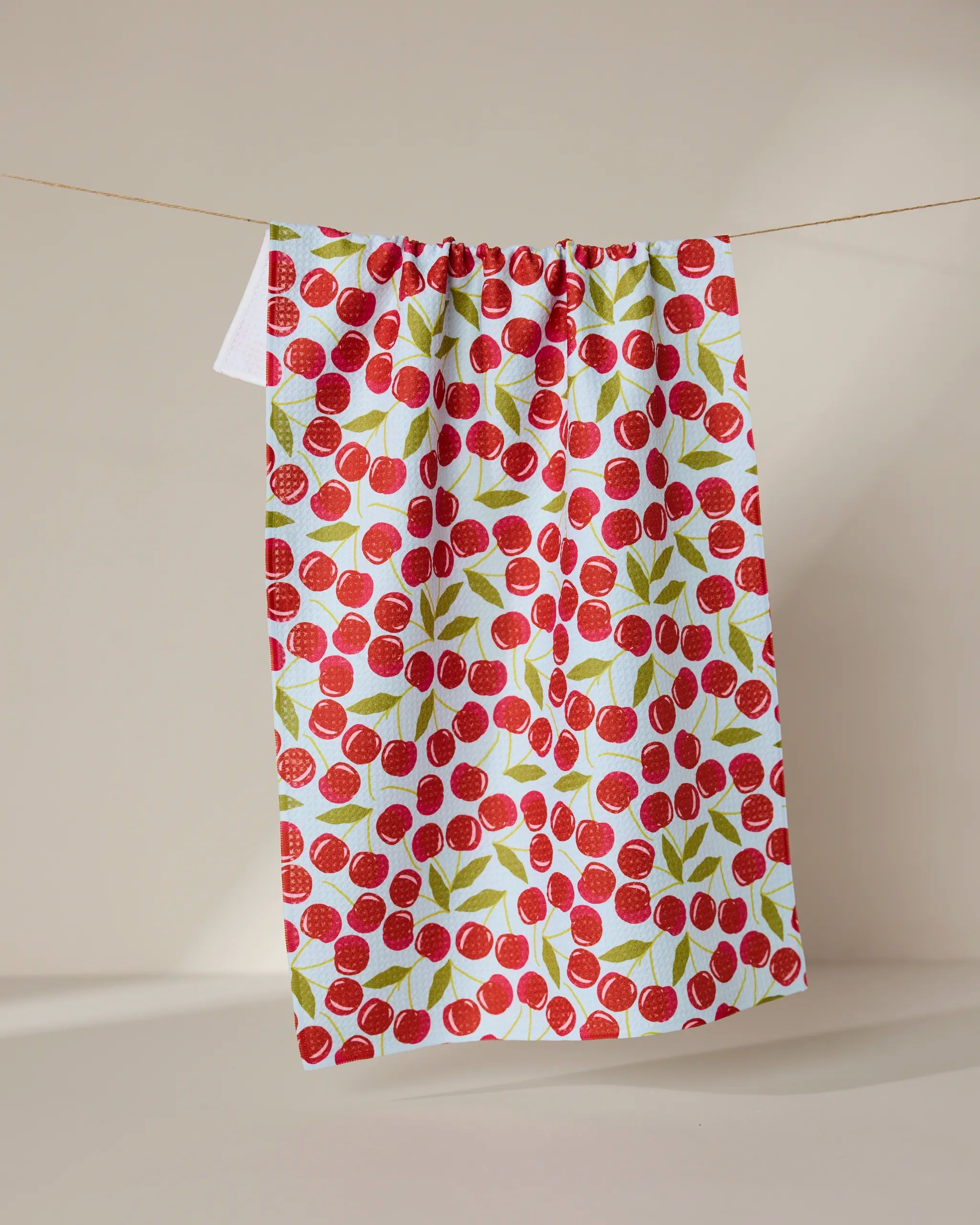 The GEOMETRY Sweet Cherry quick-drying tea towel, featuring a red cherry and green leaf pattern, hangs on a thin string against a plain beige background.