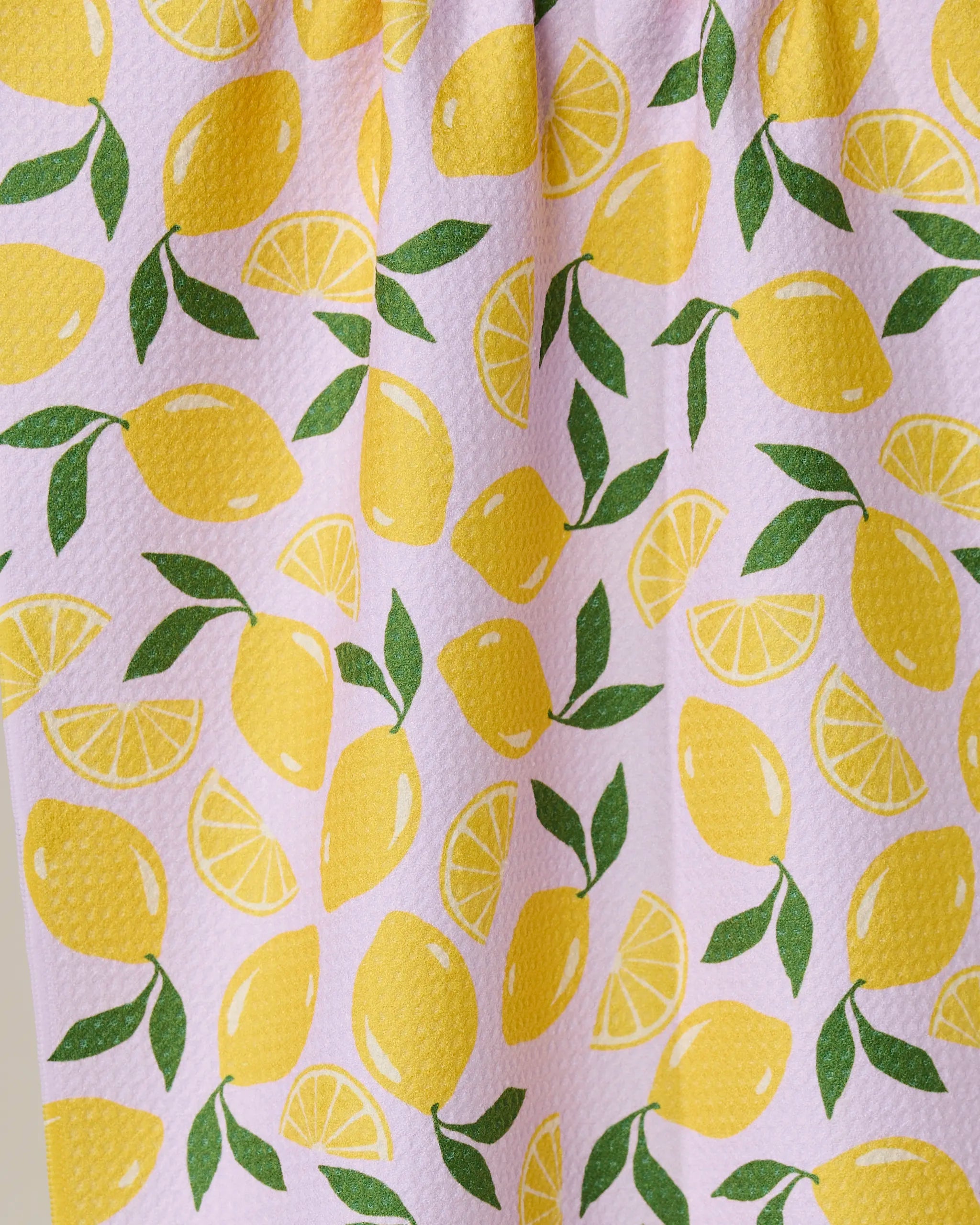 The GEOMETRY Sweet Lemon tea towel features whole yellow lemons, lemon slices, and green leaves on a light pink background. This charming, absorbent dish towel with a textured surface adds style and function to your kitchen.