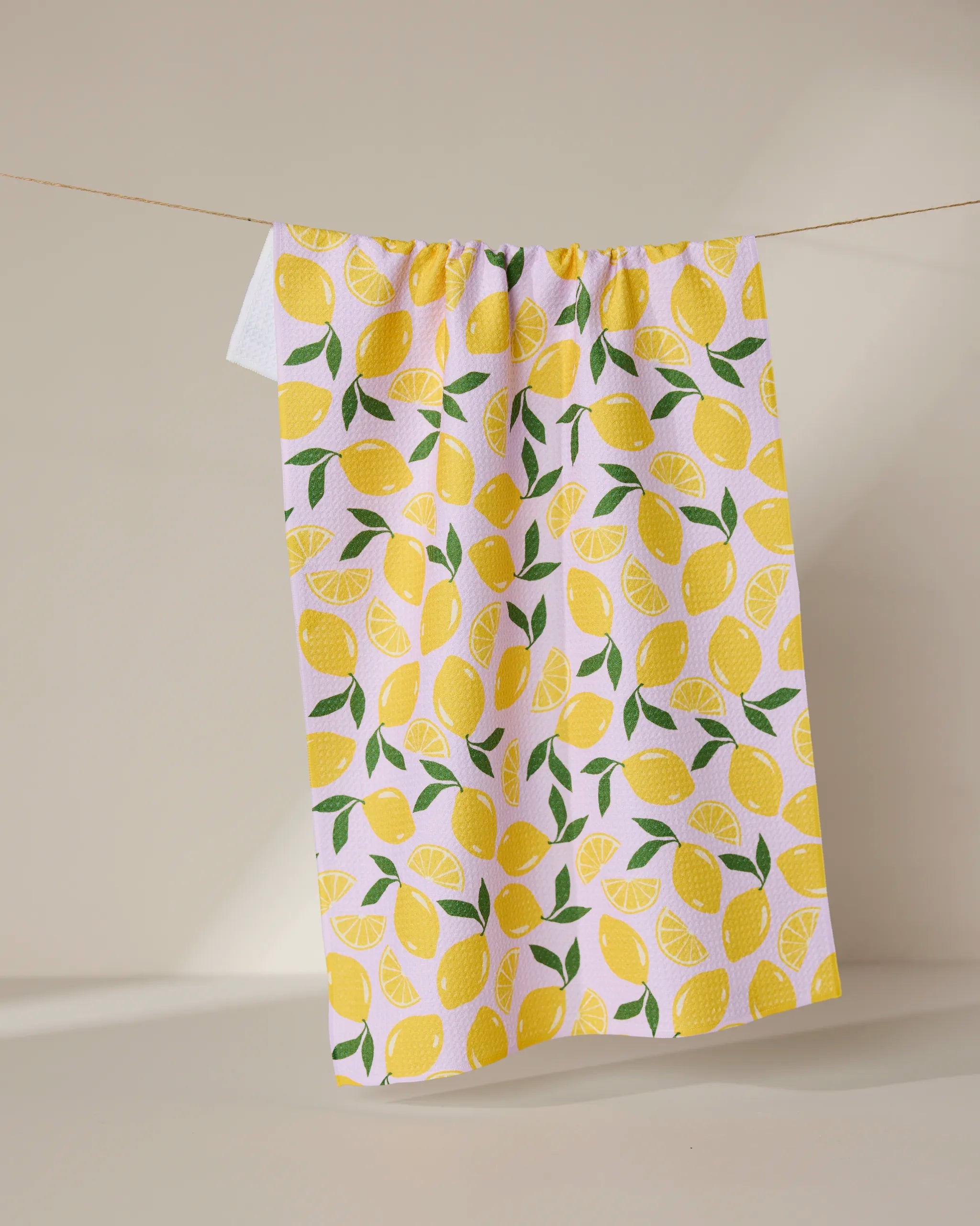 The GEOMETRY Sweet Lemon kitchen tea towel, featuring a vibrant design of whole lemons, slices, and green leaves on a white background, hangs on a string against a neutral backdrop.