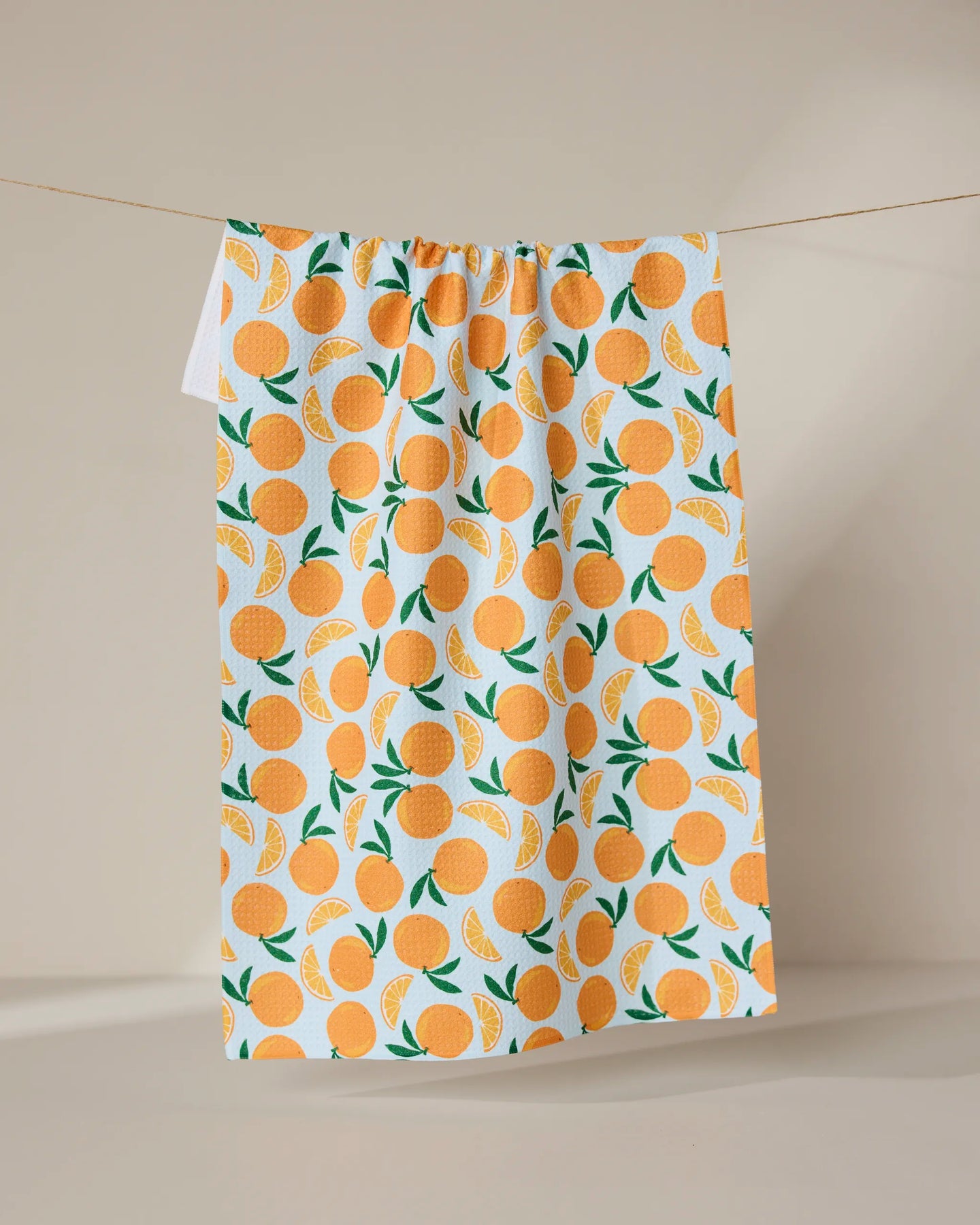 The GEOMETRY Sweet Orange tea towel features a vibrant orange fruit and green leaf pattern on white, hanging against a neutral backdrop—a stylish, quick-drying addition to any kitchen.