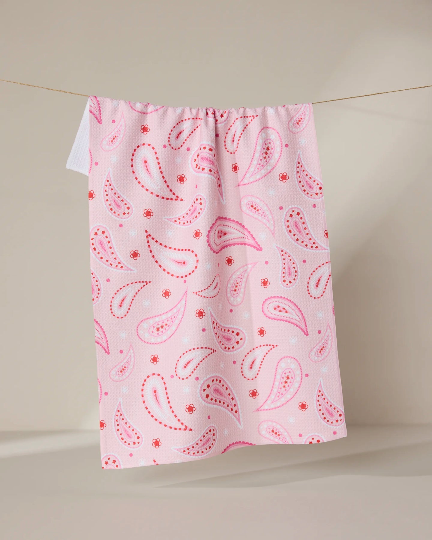 The Sweet Paisley tea towel by GEOMETRY, featuring a pink and red paisley pattern on quick-drying recycled fabric, hangs on a clothesline against a neutral beige background.