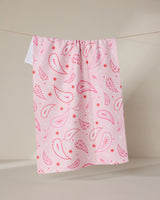 The Sweet Paisley tea towel by GEOMETRY, featuring a pink and red paisley pattern on quick-drying recycled fabric, hangs on a clothesline against a neutral beige background.