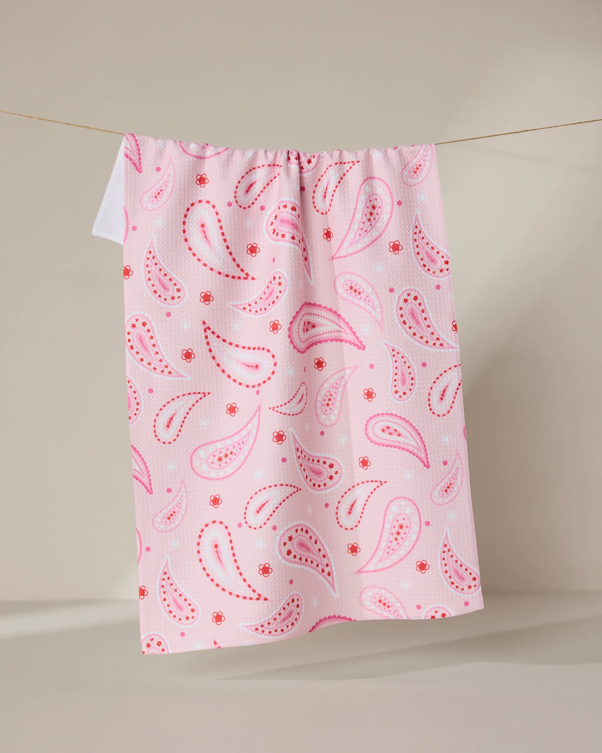 The Sweet Paisley tea towel by GEOMETRY, featuring a pink and red paisley pattern on quick-drying recycled fabric, hangs on a clothesline against a neutral beige background.