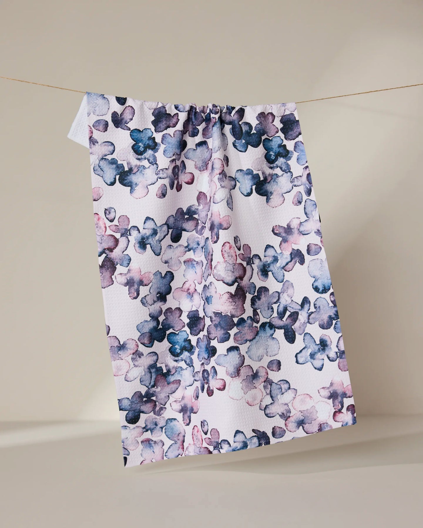 The Sweet Petals tea towel by GEOMETRY, made from recycled materials, displays a blue and purple watercolor floral pattern and hangs from a clothesline against a neutral background.