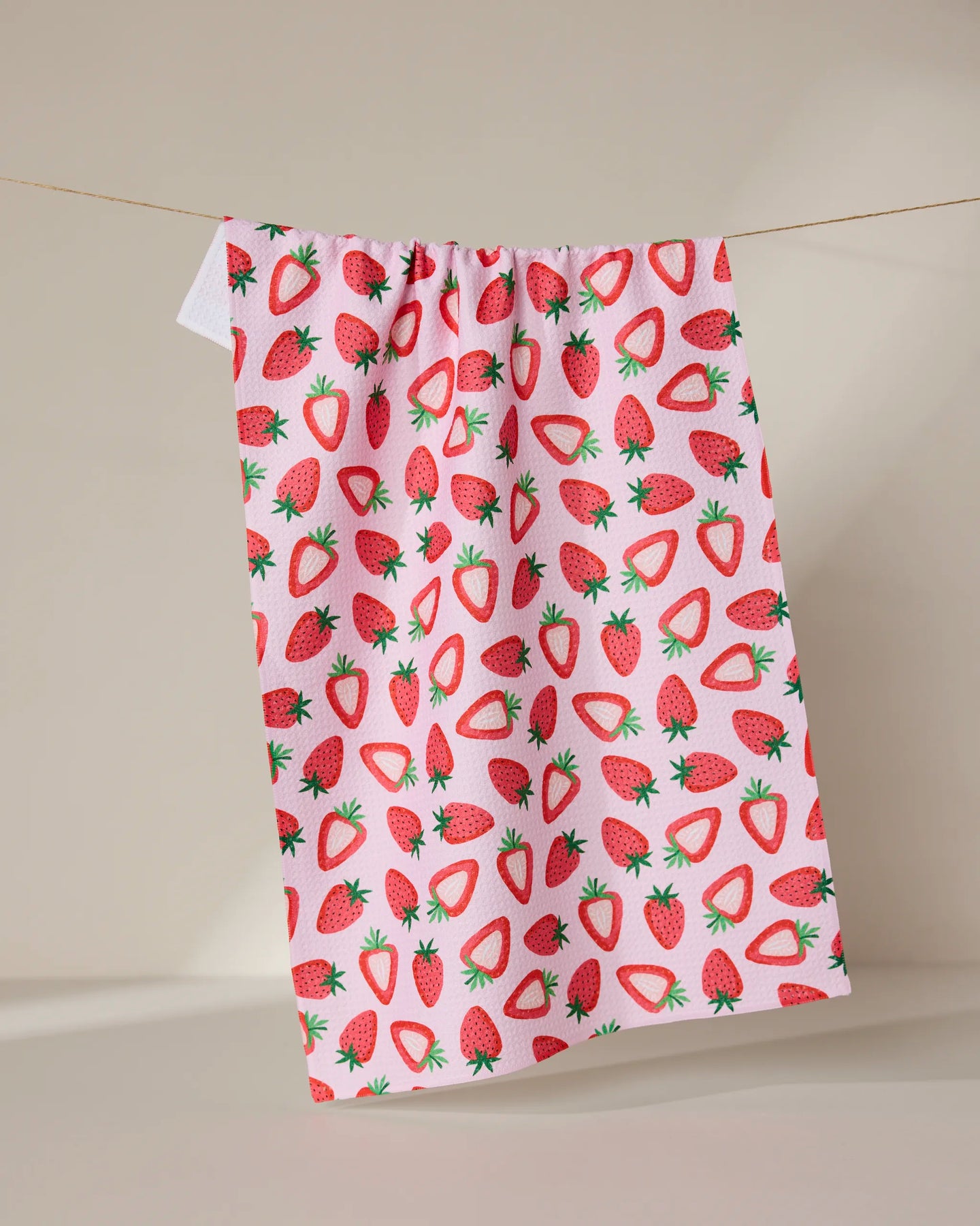 The GEOMETRY Sweet Strawberry kitchen towel, ultra-absorbent and featuring a colorful whole and sliced strawberry pattern on white, hangs from a clothesline against a plain beige background.