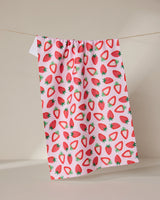 The GEOMETRY Sweet Strawberry kitchen towel, ultra-absorbent and featuring a colorful whole and sliced strawberry pattern on white, hangs from a clothesline against a plain beige background.