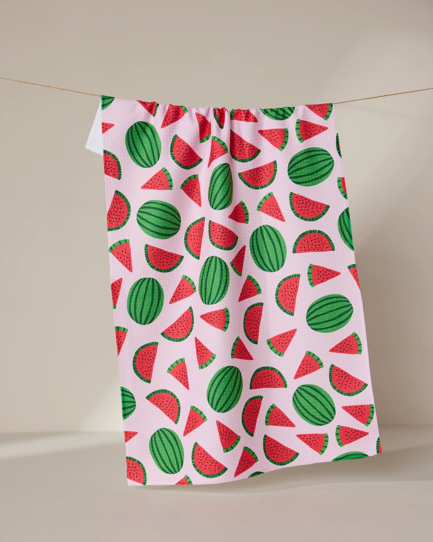 The GEOMETRY Sweet Watermelon tea towel, featuring colorful watermelon slice and whole fruit designs on white, hangs on a string against a neutral background.
