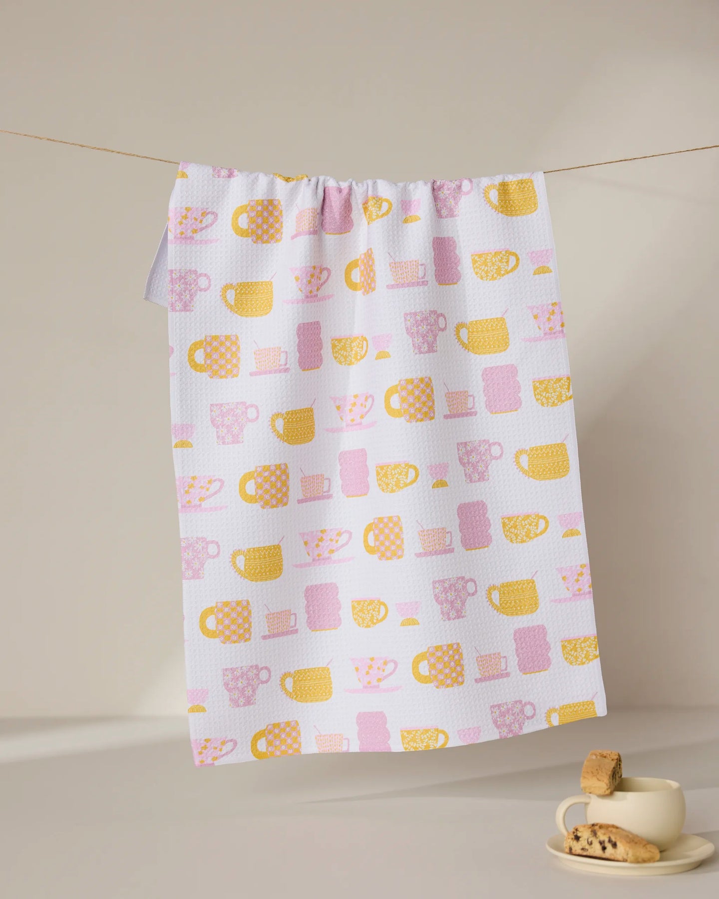The GEOMETRY Talk Over Tea towel, made from recycled materials and featuring yellow and pink mugs and cups, hangs on a line above a beige cup and saucer with cookies, set on a light surface against a neutral background.