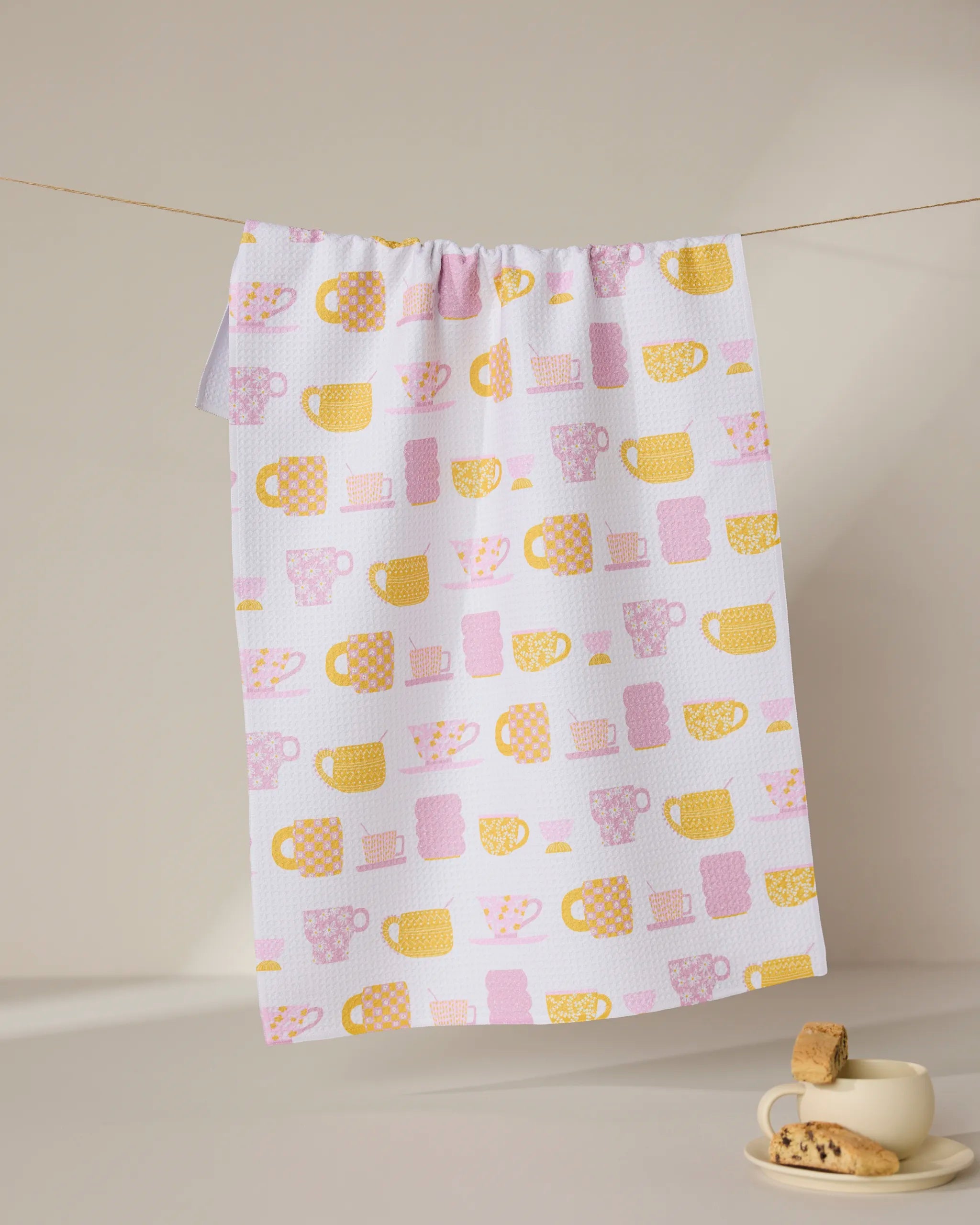 The GEOMETRY Talk Over Tea towel, made from recycled materials and featuring yellow and pink mugs and cups, hangs on a line above a beige cup and saucer with cookies, set on a light surface against a neutral background.