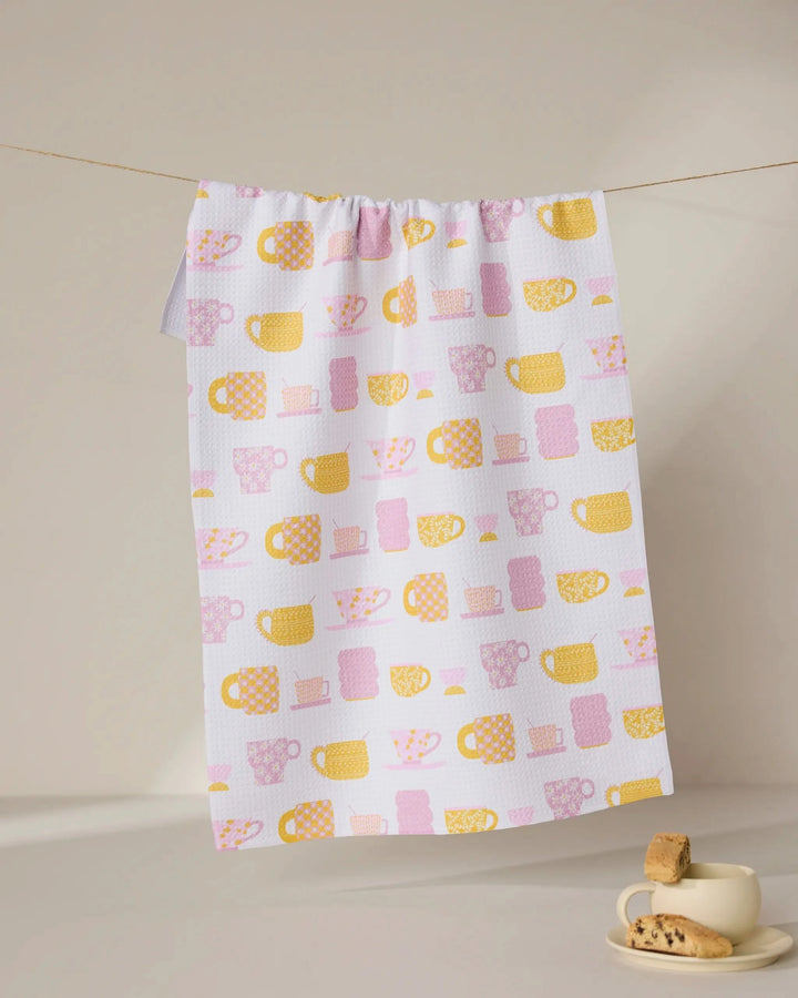 The GEOMETRY Talk Over Tea towel, made from recycled materials and featuring yellow and pink mugs and cups, hangs on a line above a beige cup and saucer with cookies, set on a light surface against a neutral background.
