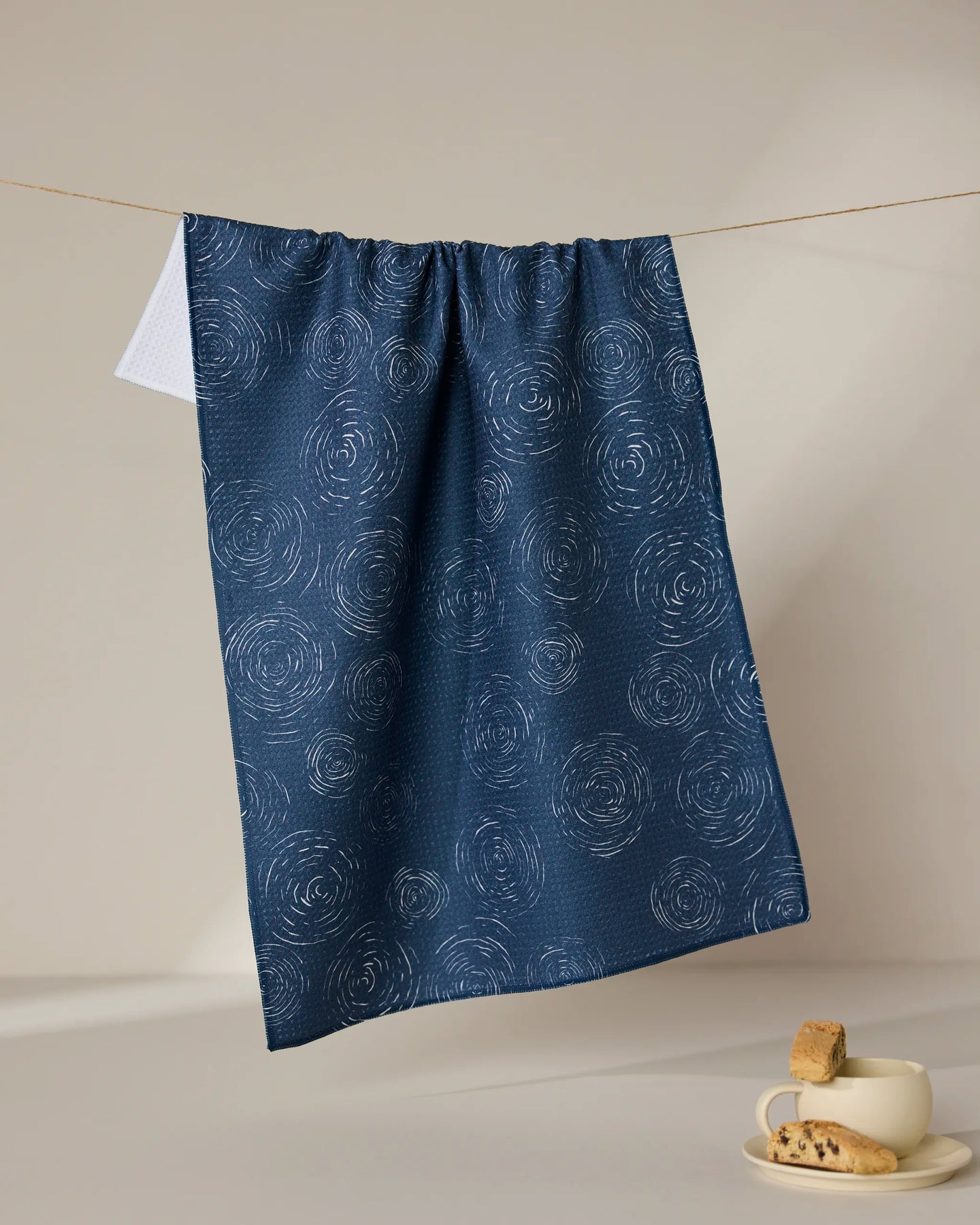 The GEOMETRY Taps - Midnight recycled navy blue kitchen towel with a white swirl pattern hangs on a string. Below, a cup with a drink and cookies rests on a beige surface against a neutral background.