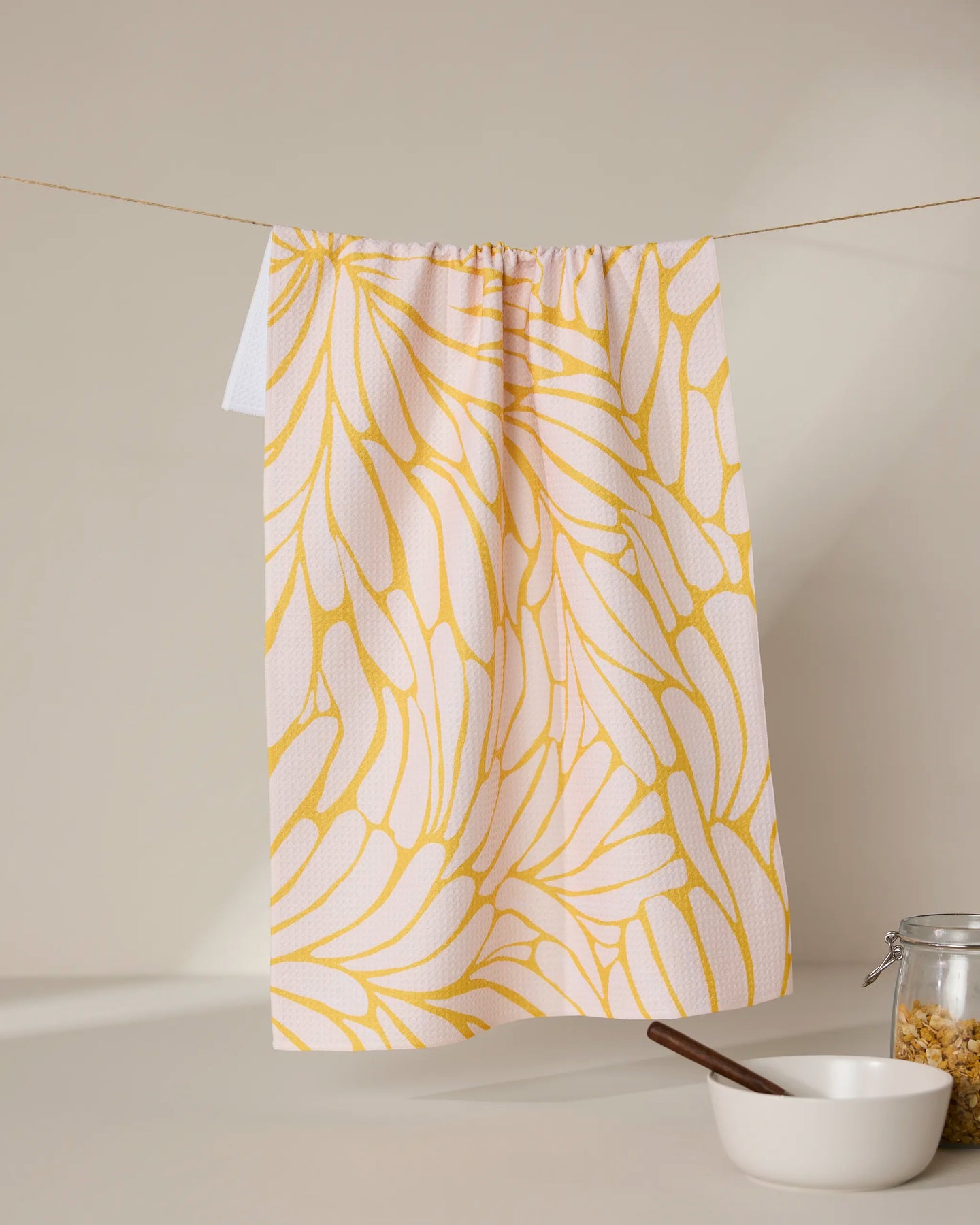 The GEOMETRY Tenderhearted tea towel, featuring a yellow abstract floral pattern, hangs on a string above a white bowl with a wooden spoon and a glass jar of cereal on a beige surface against a neutral background.