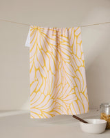 The GEOMETRY Tenderhearted tea towel, featuring a yellow abstract floral pattern, hangs on a string above a white bowl with a wooden spoon and a glass jar of cereal on a beige surface against a neutral background.
