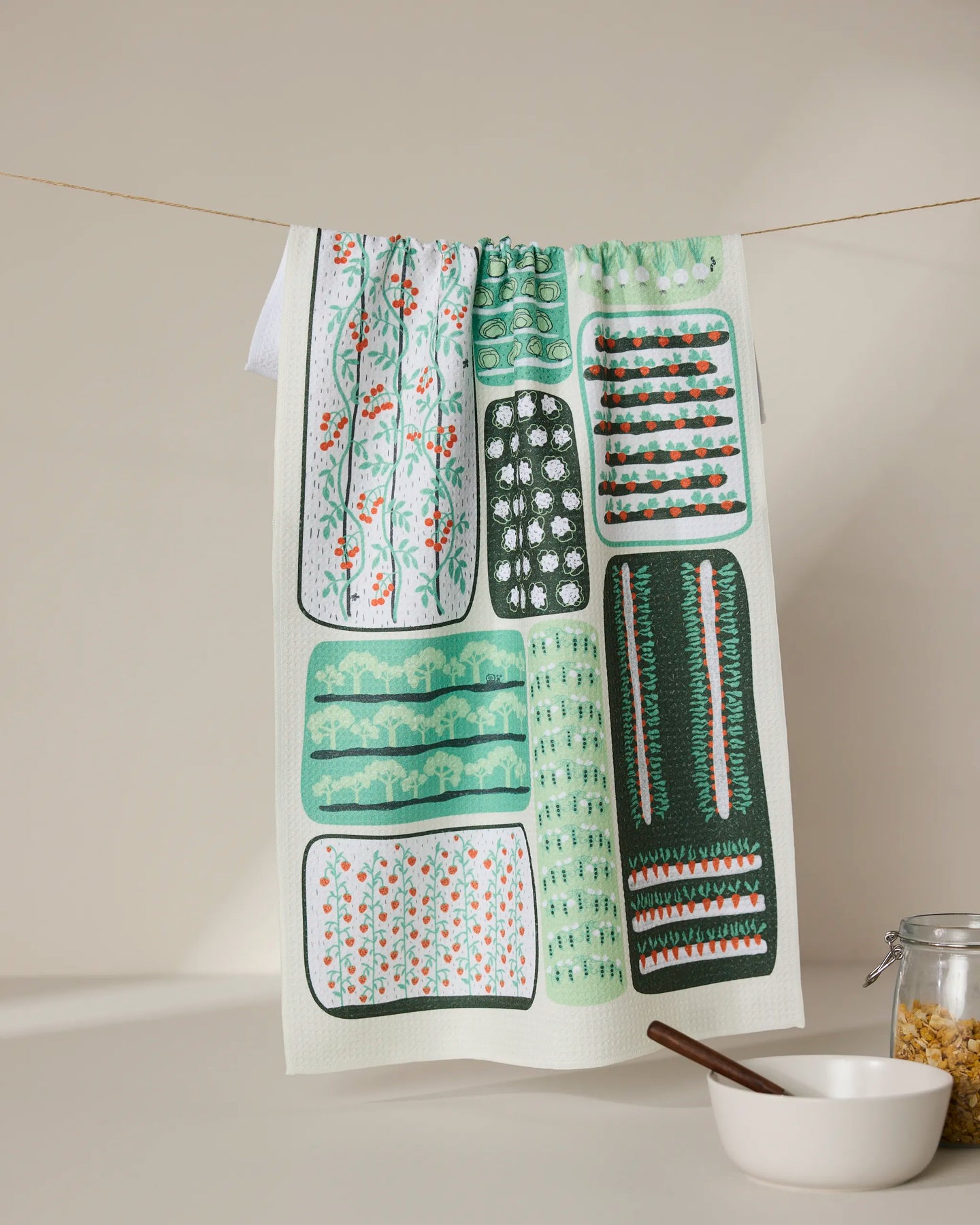 The GEOMETRY Tending tea towel, featuring quick-dry fabric and green, teal, and orange garden-inspired patterns, hangs on a line above a white bowl with a wooden spoon and a glass jar of pasta against a beige background.
