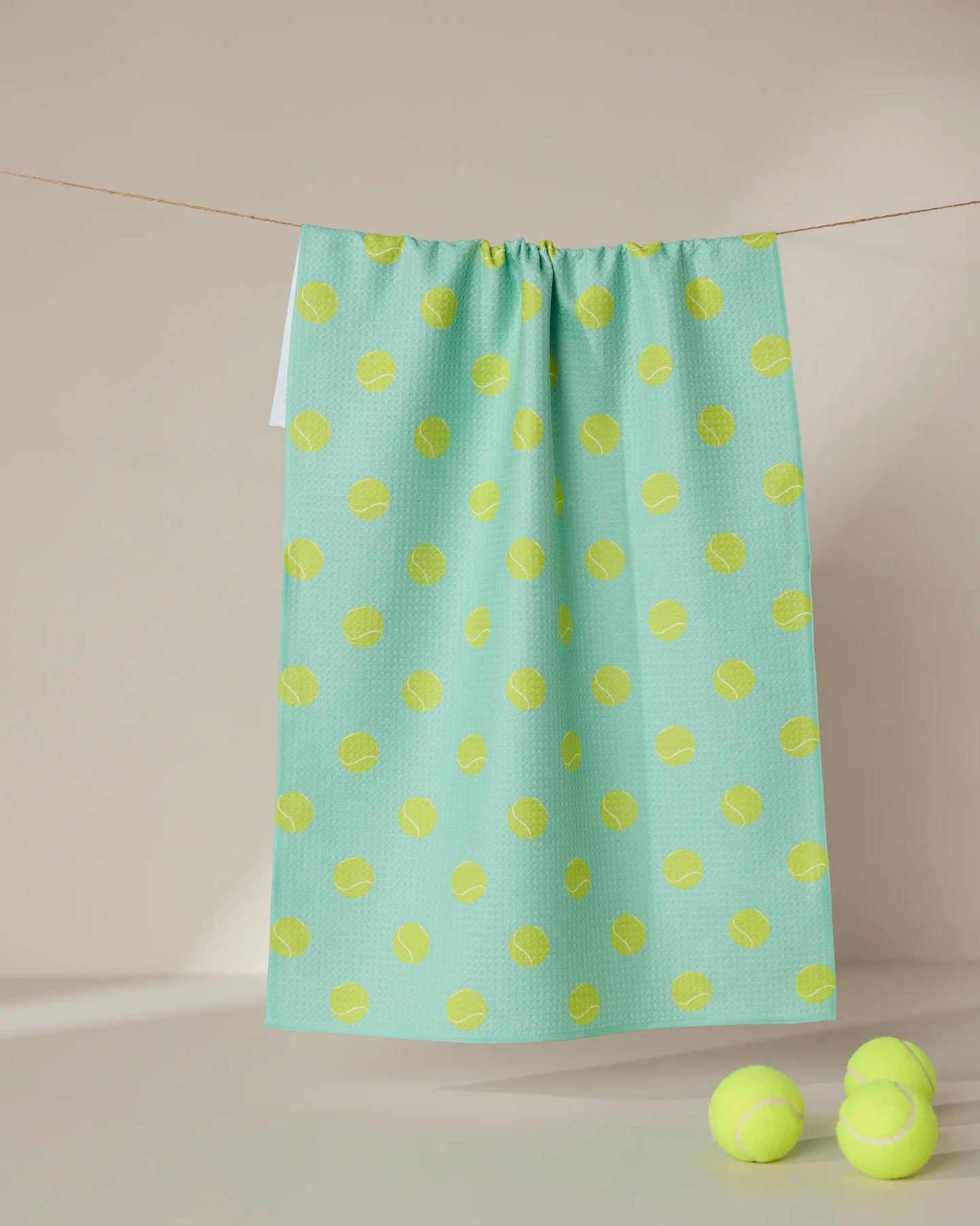 A GEOMETRY Tennis Ball hangs in a light green, ultra-absorbent towel with a yellow tennis ball pattern on a clothesline. Three yellow GEOMETRY Tennis Balls lie nearby, all set against a minimal beige background.