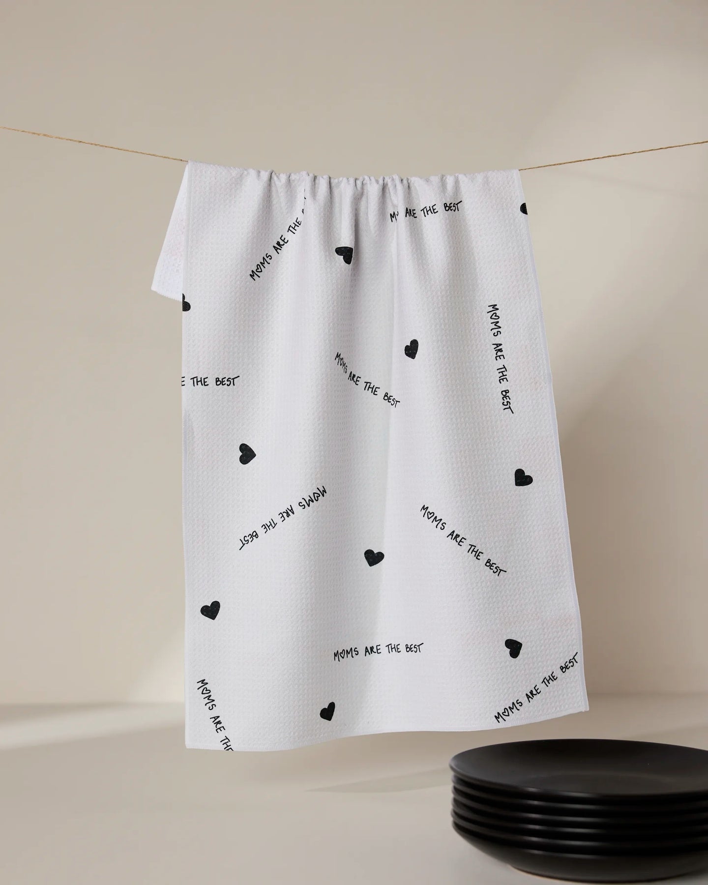 A white ultra-absorbent tea towel by GEOMETRY, called The Best, featuring small black hearts and MOMS ARE THE BEST print, hangs above black plates.