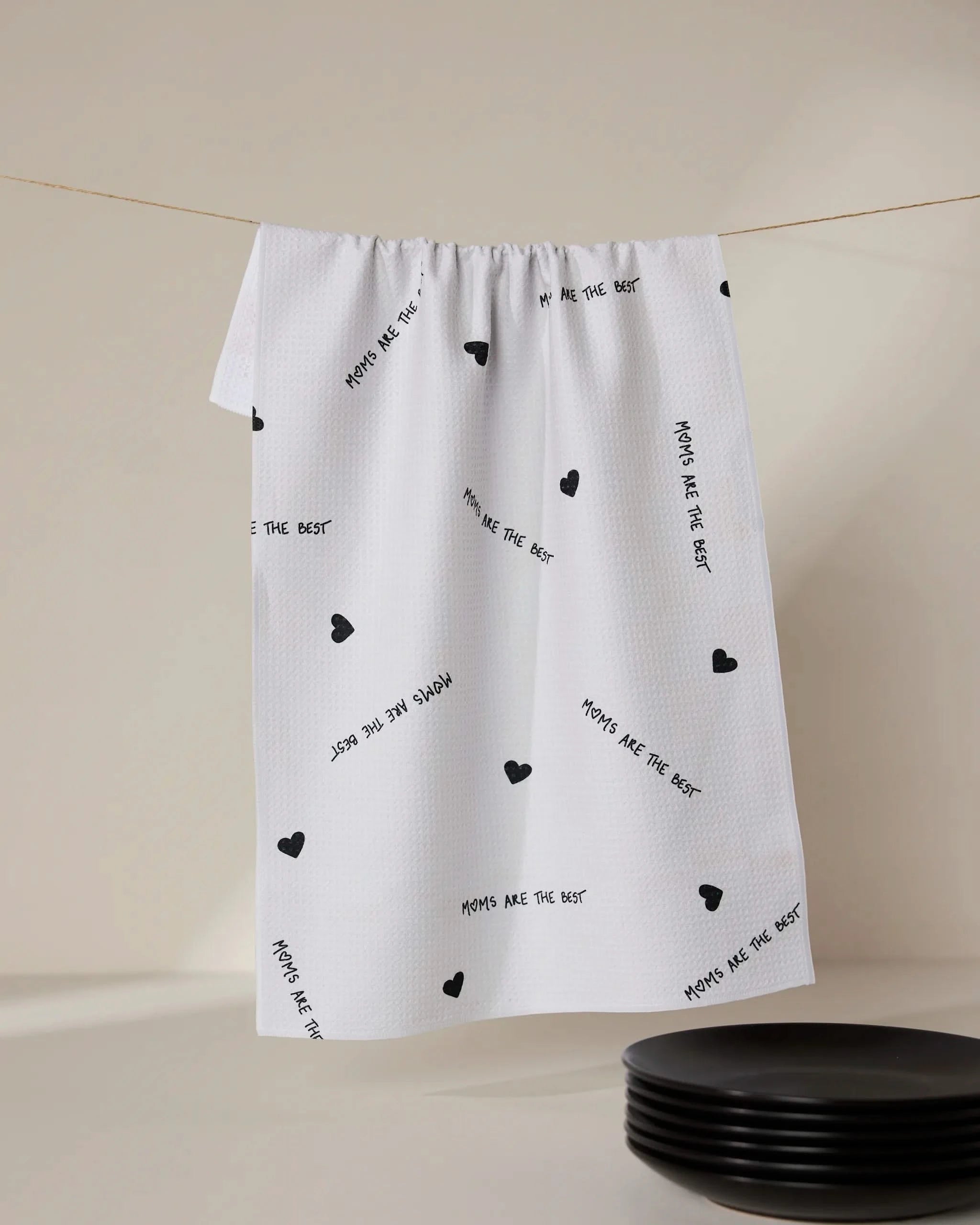 A white ultra-absorbent tea towel by GEOMETRY, called The Best, featuring small black hearts and MOMS ARE THE BEST print, hangs above black plates.