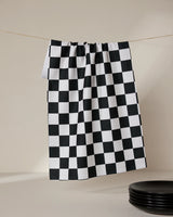 The Classic tea towel by GEOMETRY, made from recycled materials in a black and white checkered pattern, hangs from a clothesline against a neutral background with black plates stacked in the lower right corner.