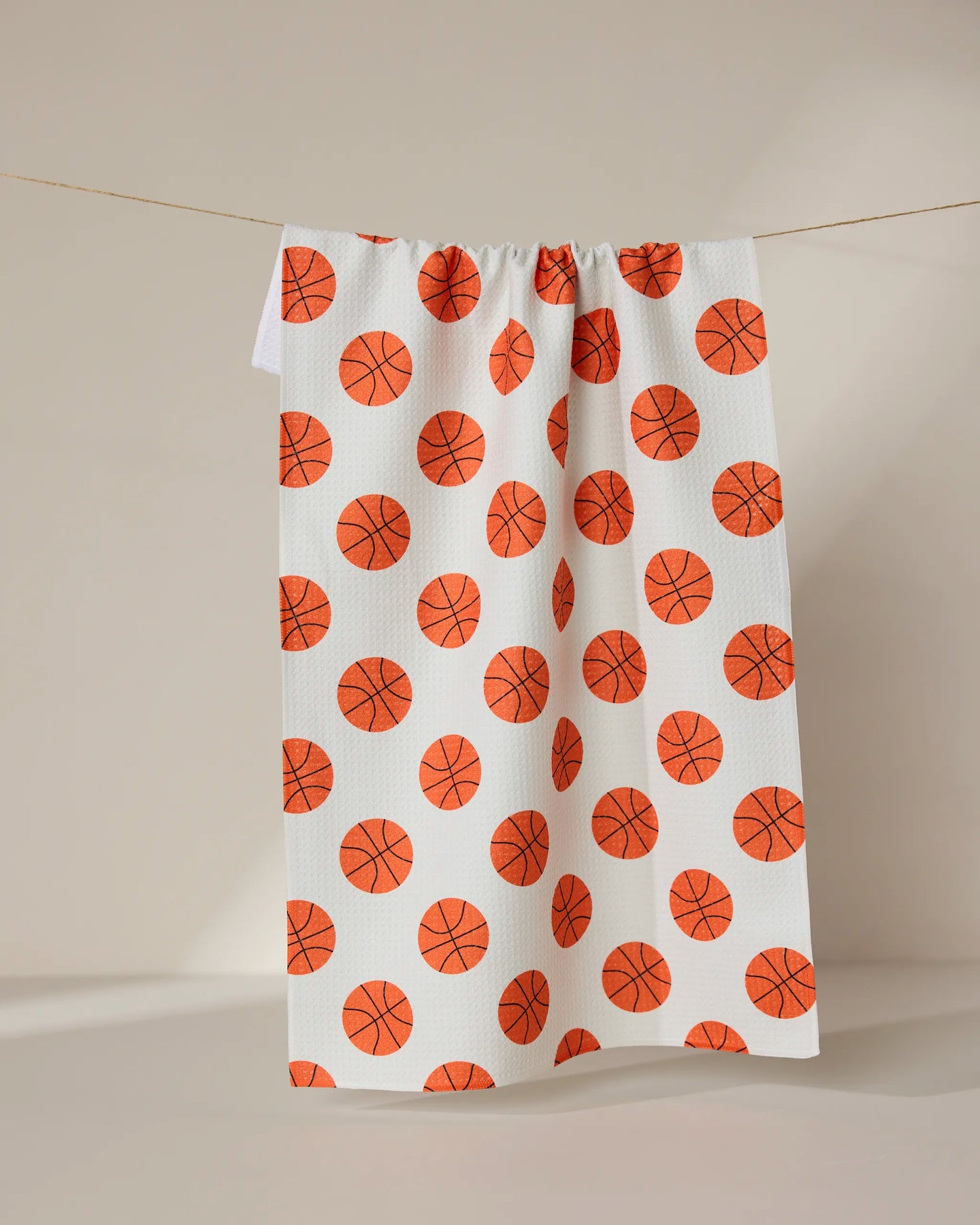 The Three Pointer tea towel by GEOMETRY, featuring an all-over orange basketball pattern and made from recycled materials, hangs on a clothesline against a neutral background.