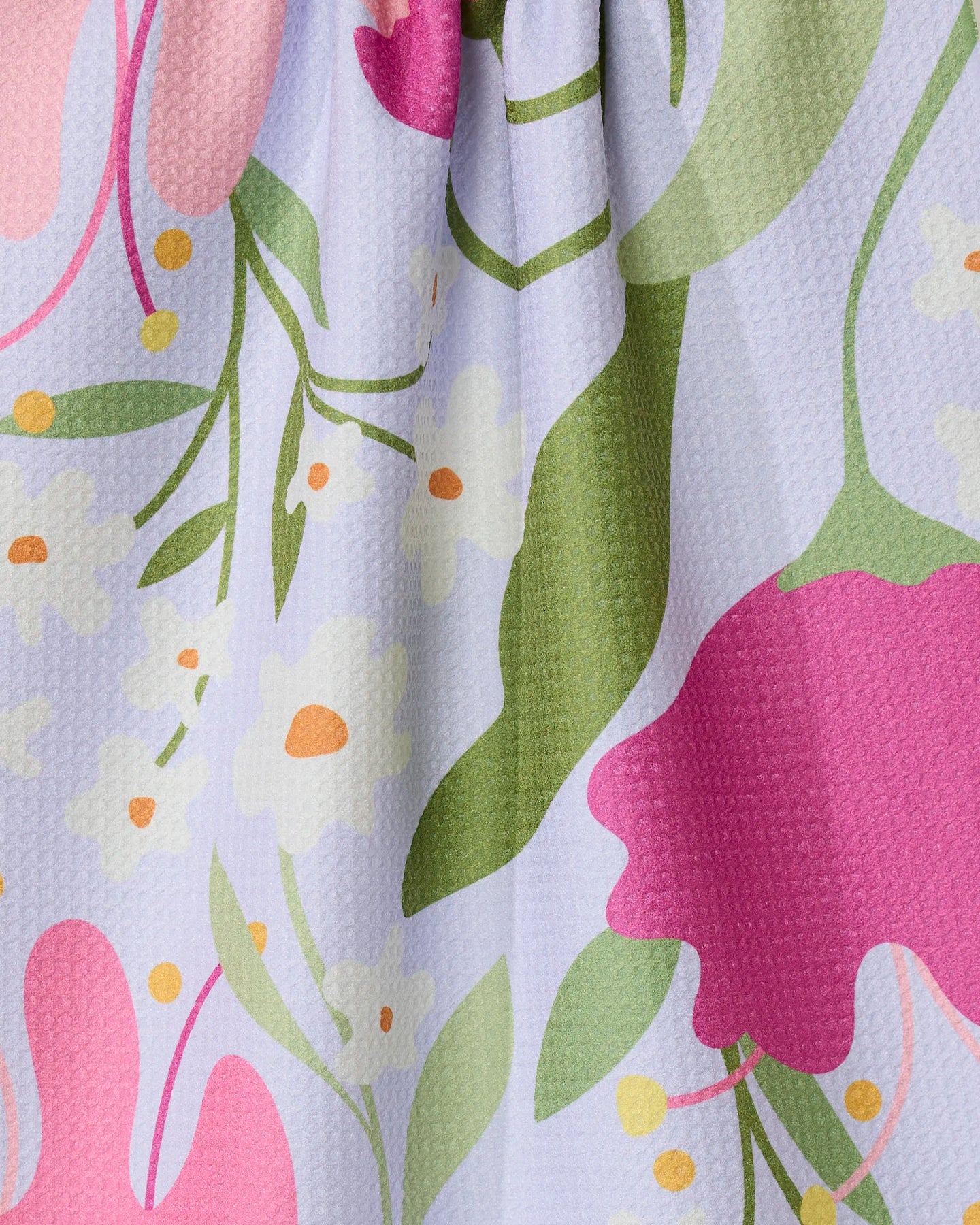 A close-up of the GEOMETRY Thoughtful tea towel, made from recycled materials, shows a colorful floral pattern with large pink flowers, green leaves, yellow dots, and small white blossoms on a textured light purple background.