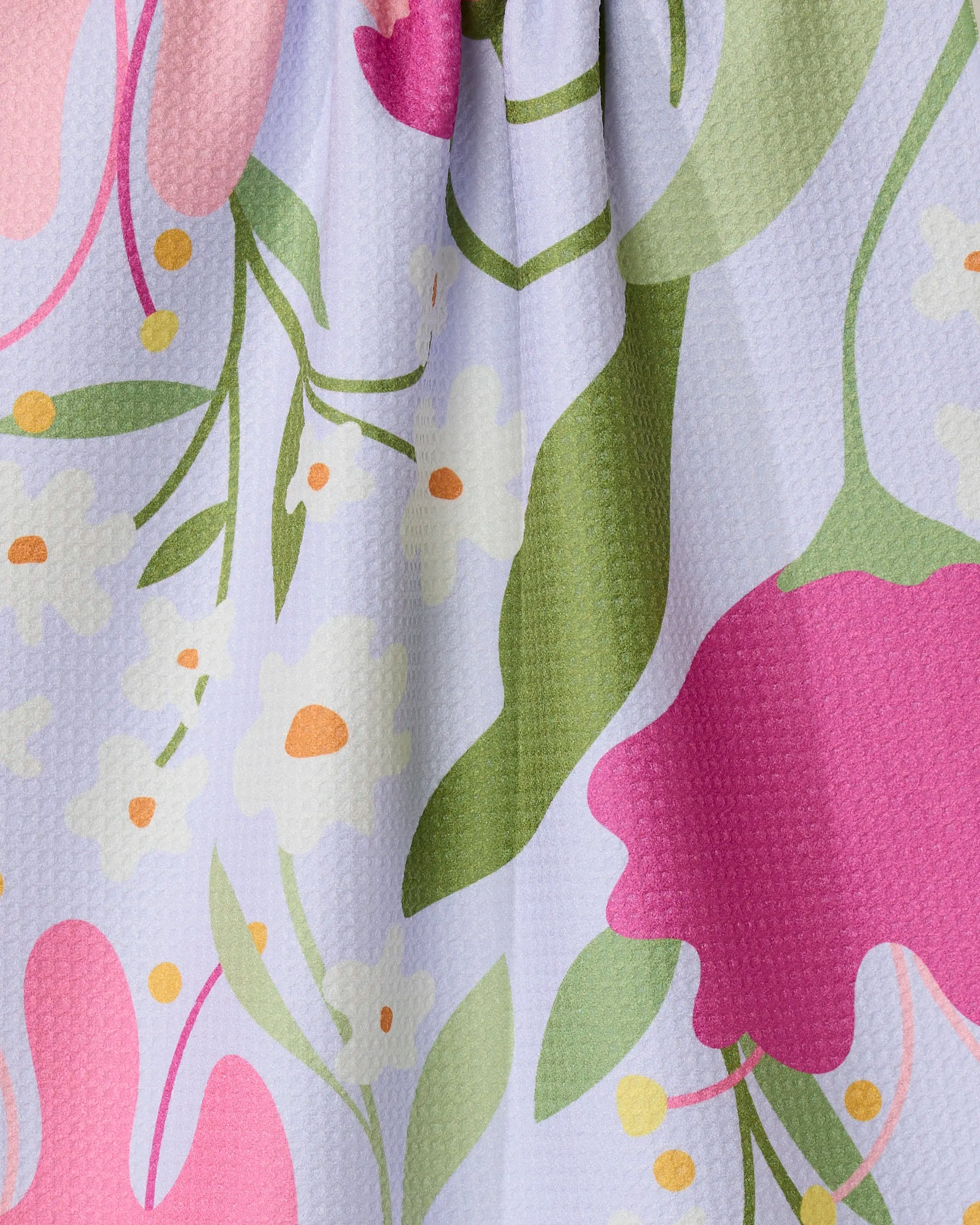 A close-up of the GEOMETRY Thoughtful tea towel, made from recycled materials, shows a colorful floral pattern with large pink flowers, green leaves, yellow dots, and small white blossoms on a textured light purple background.