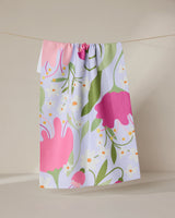 The GEOMETRY Thoughtful tea towel, made from recycled materials, features a vibrant floral design with large pink and red flowers, green leaves, and yellow-white accents. It hangs on a beige background and dries quickly.