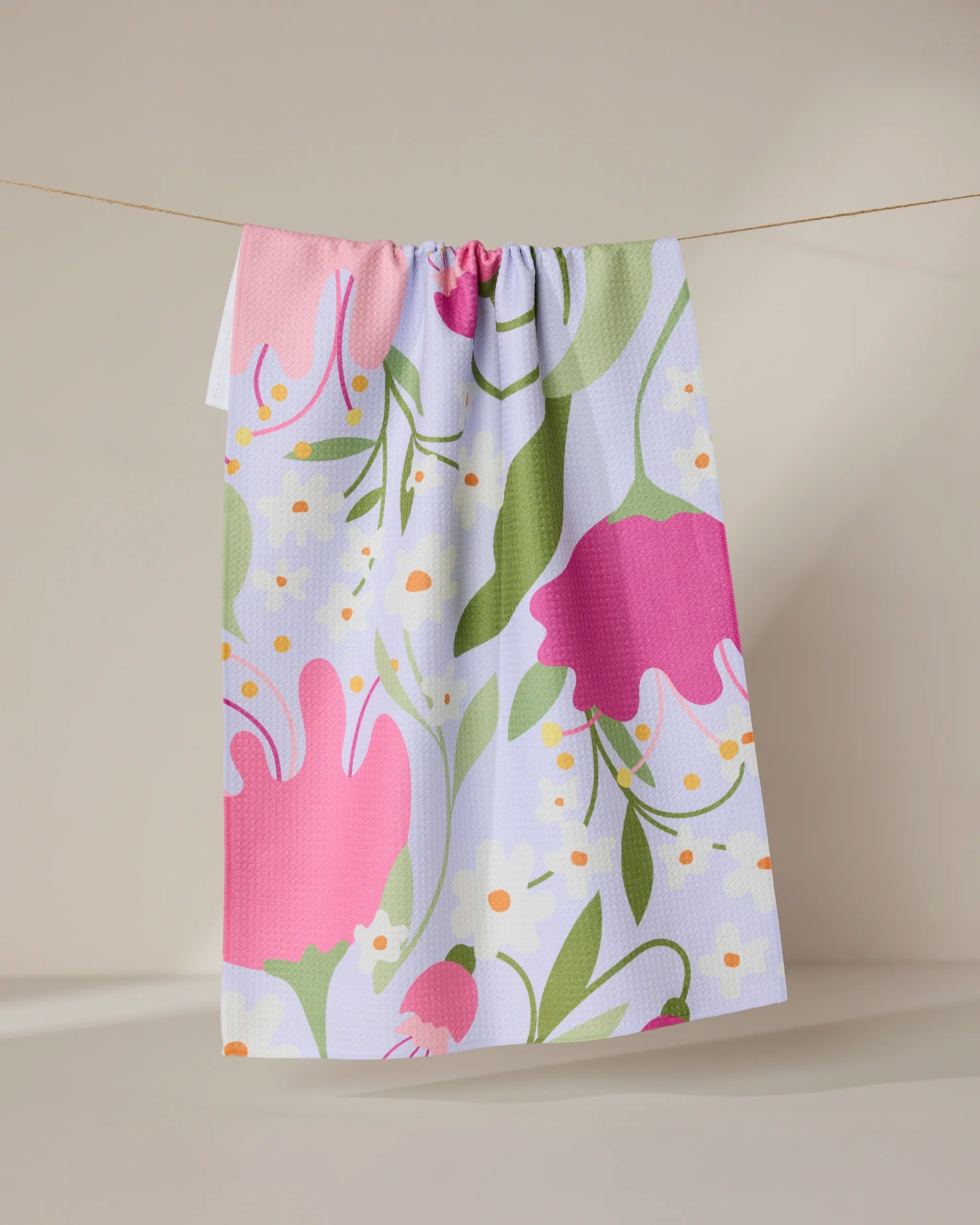 The GEOMETRY Thoughtful tea towel, made from recycled materials, features a vibrant floral design with large pink and red flowers, green leaves, and yellow-white accents. It hangs on a beige background and dries quickly.