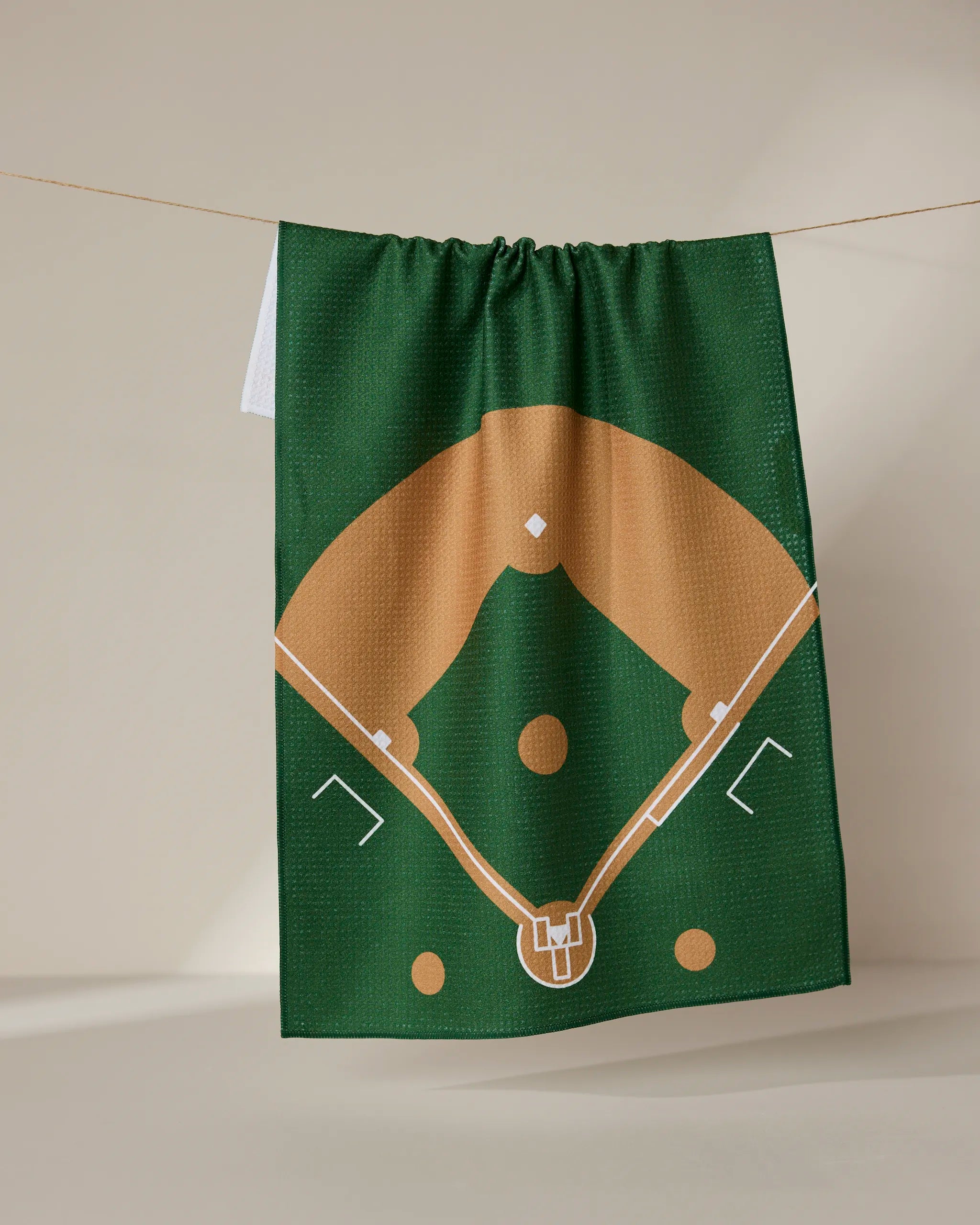 The Field tea towel by GEOMETRY, made from recycled materials, displays a brown and white baseball diamond illustration on green fabric and hangs against a neutral background.
