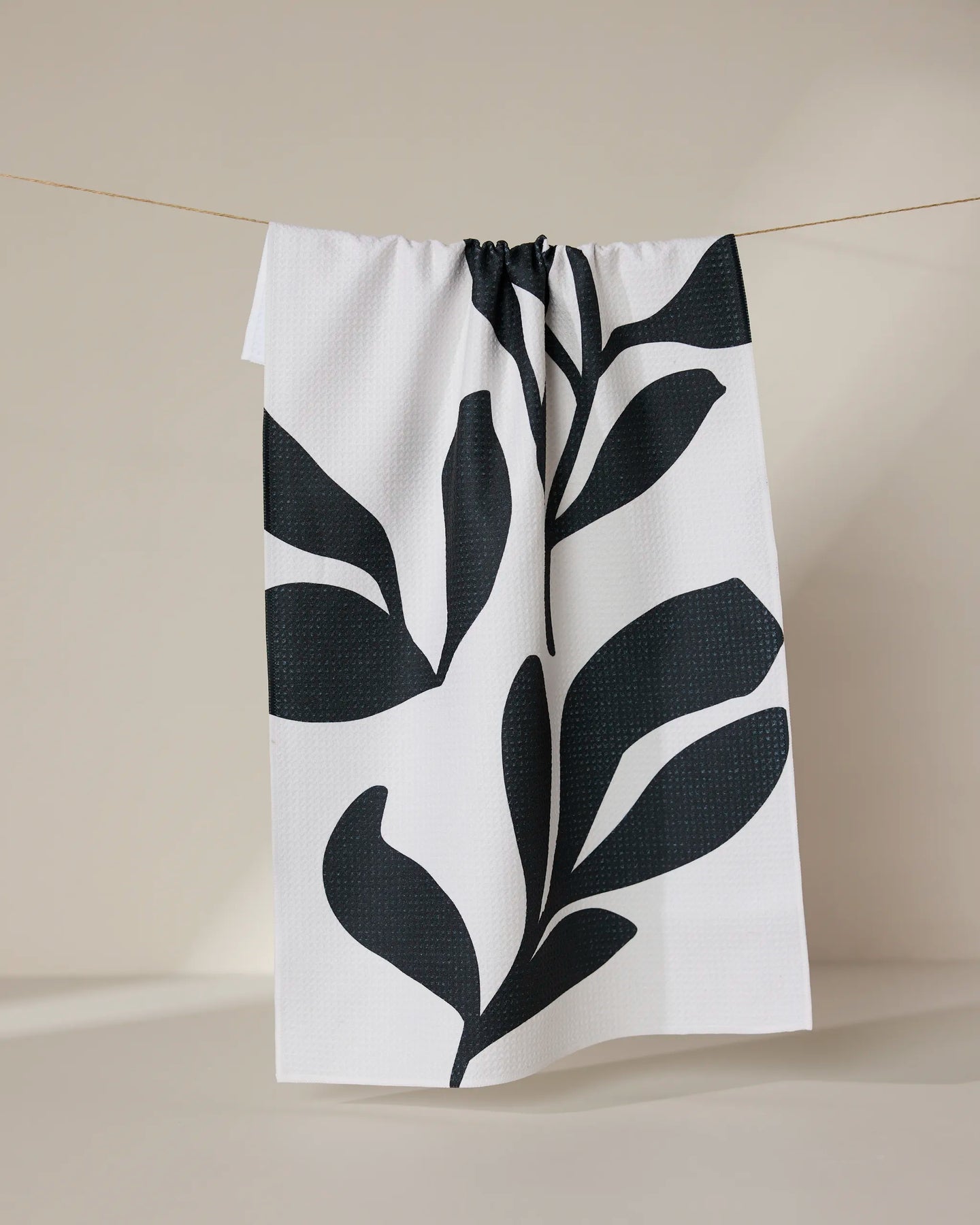 The GEOMETRY Threes tea towel, made from recycled materials, features large black abstract leaf patterns on white and hangs on a string against a plain, neutral background.