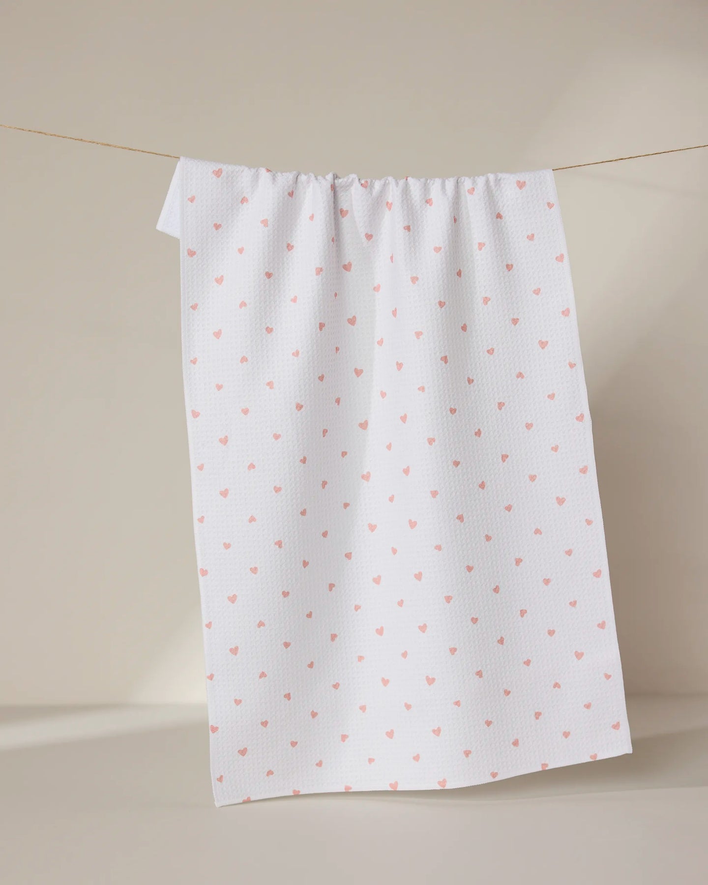 The GEOMETRY Tiny Hearts towel, ultra-absorbent and white with a pattern of small pink hearts, hangs on a clothesline against a neutral beige background.