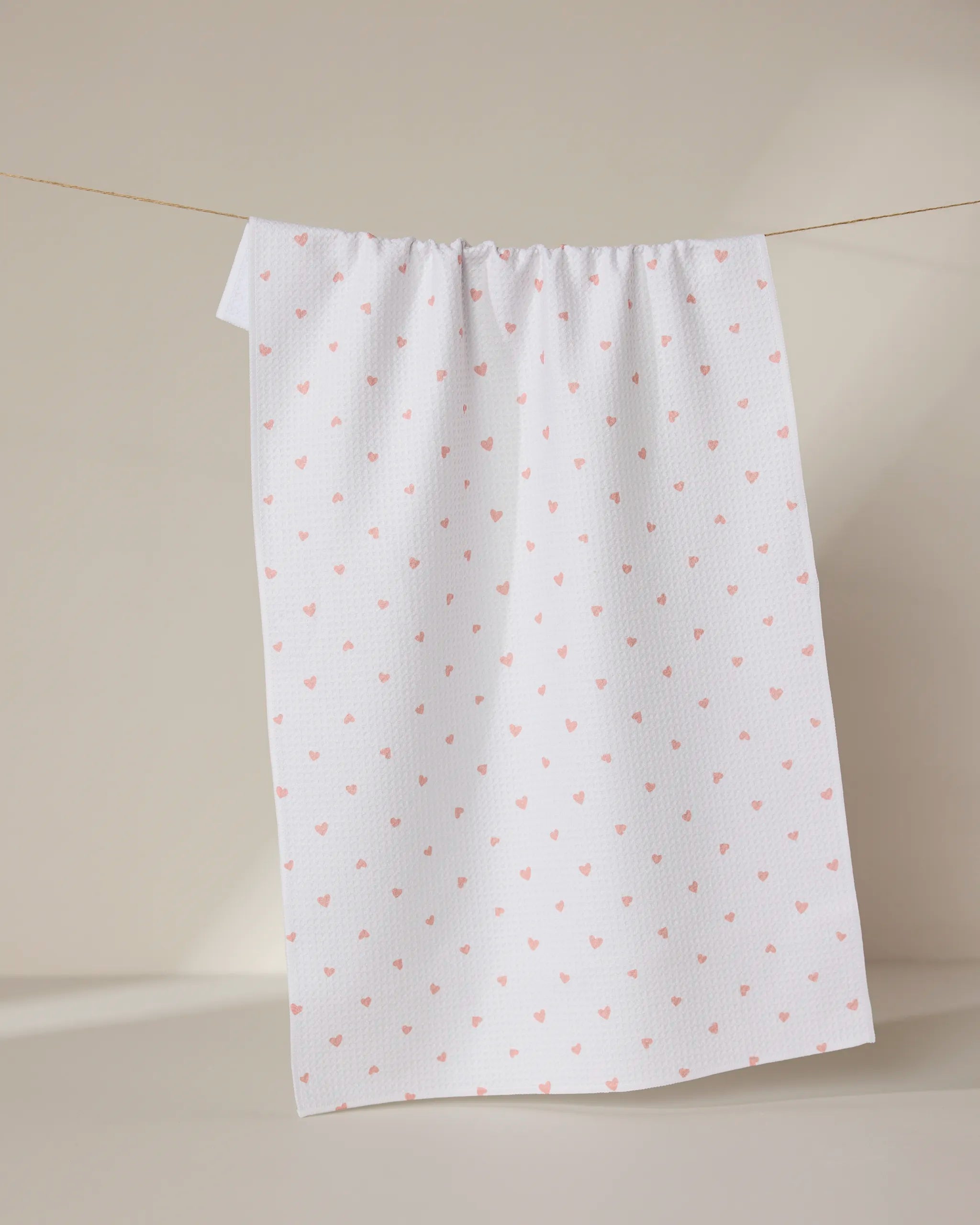 The GEOMETRY Tiny Hearts towel, ultra-absorbent and white with a pattern of small pink hearts, hangs on a clothesline against a neutral beige background.