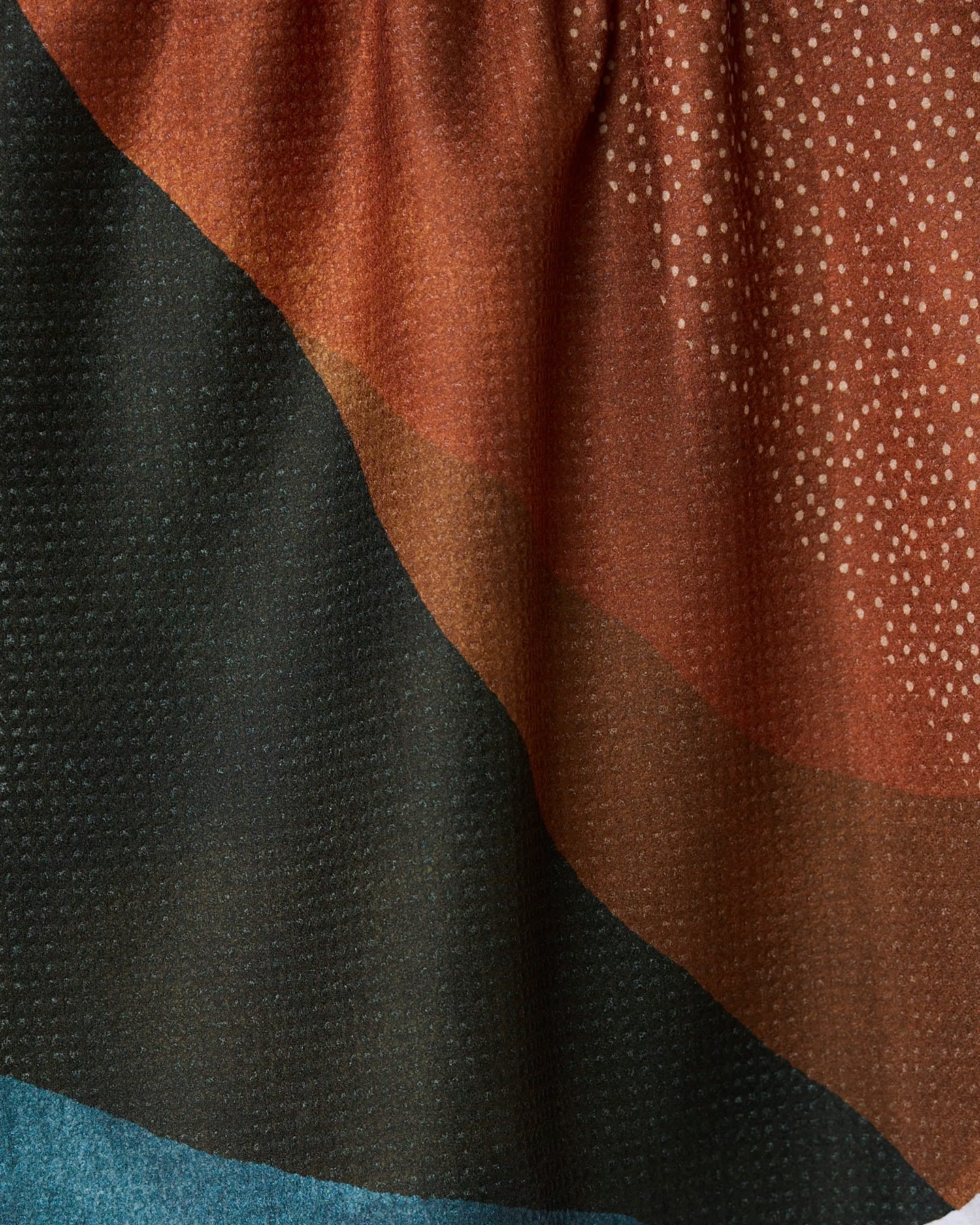Close-up of GEOMETRYs Tres kitchen towel, showcasing its textured recycled fabric with wavy black, brown, and reddish-orange patterns accented by small dots and a blue corner at the bottom left.