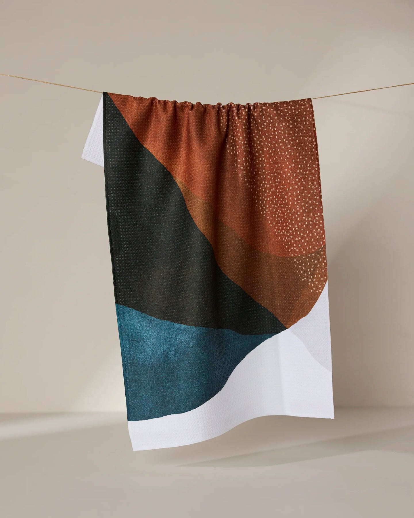 The GEOMETRY Tres recycled kitchen towel, with an abstract design in brown, black, teal, and white plus small white dots, hangs on a line against a neutral background.