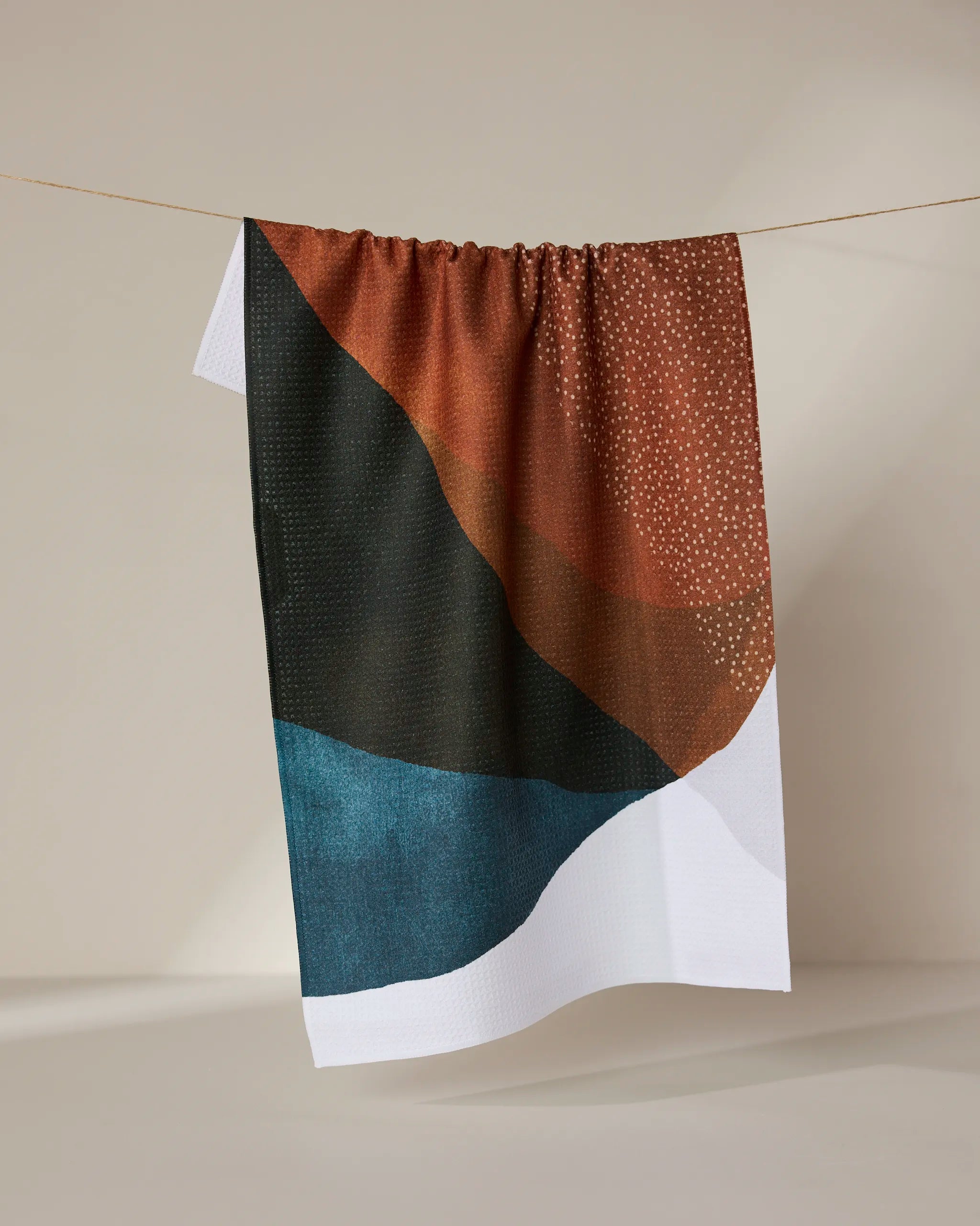 The GEOMETRY Tres recycled kitchen towel, with an abstract design in brown, black, teal, and white plus small white dots, hangs on a line against a neutral background.