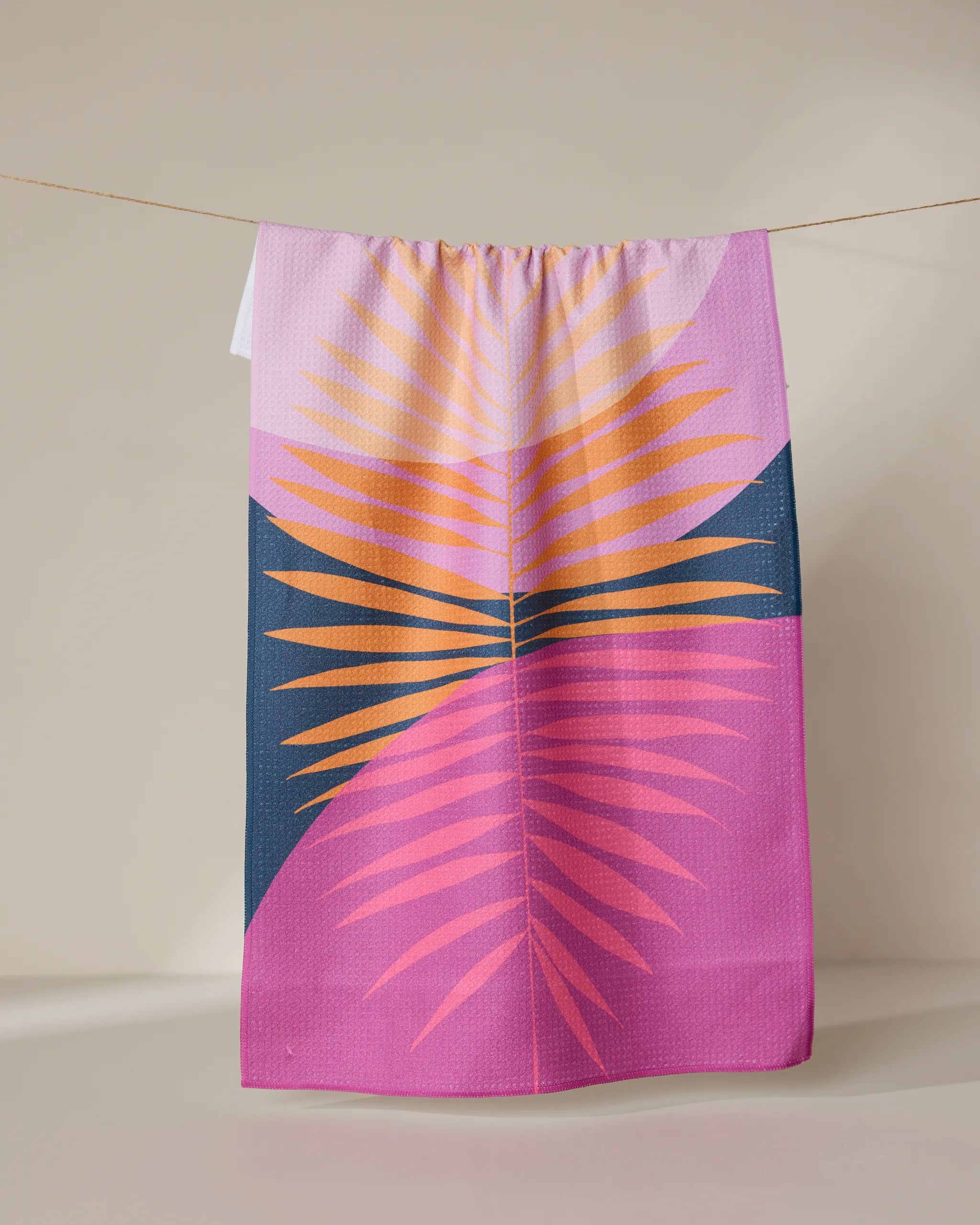 The GEOMETRY Tropical Summer tea towel, made with Geoweave fabric and featuring vibrant palm leaves on a purple and dark blue background, hangs on a clothesline against a neutral wall.