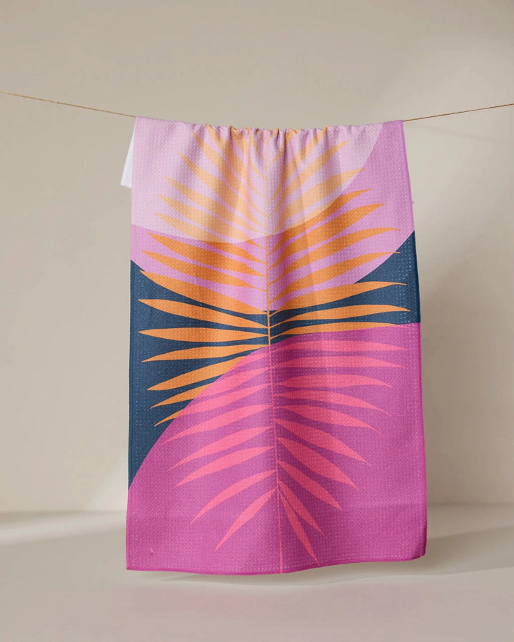The GEOMETRY Tropical Summer tea towel, made with Geoweave fabric and featuring vibrant palm leaves on a purple and dark blue background, hangs on a clothesline against a neutral wall.