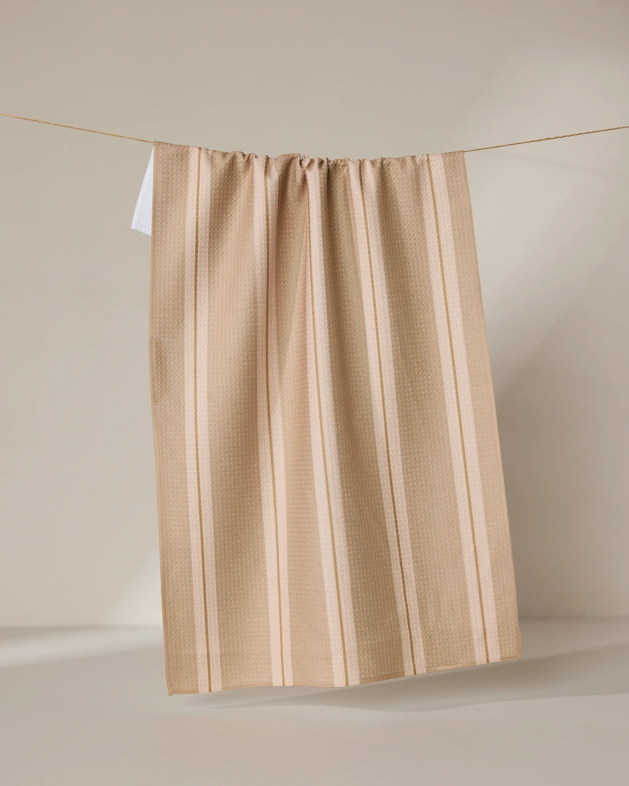 The Vanilla Chai tea towel by GEOMETRY, featuring beige and cream stripes and made from recycled materials, hangs on a clothesline against a neutral background with soft light casting gentle shadows.