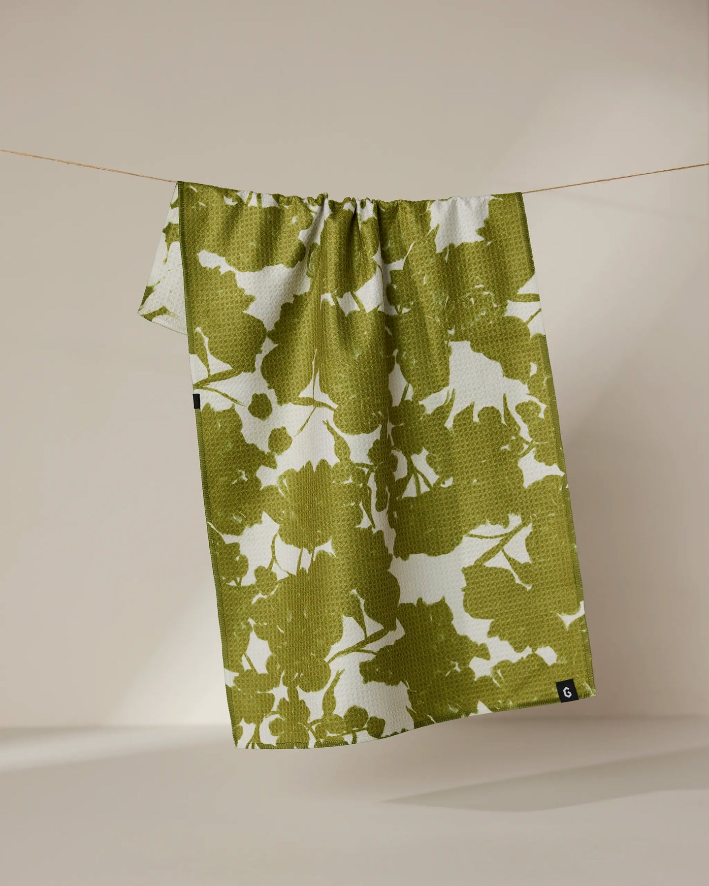 The Geometry Sweet Green tea towel, featuring a green and white abstract leaf pattern and double-sided design, hangs on a clothesline against a neutral background.