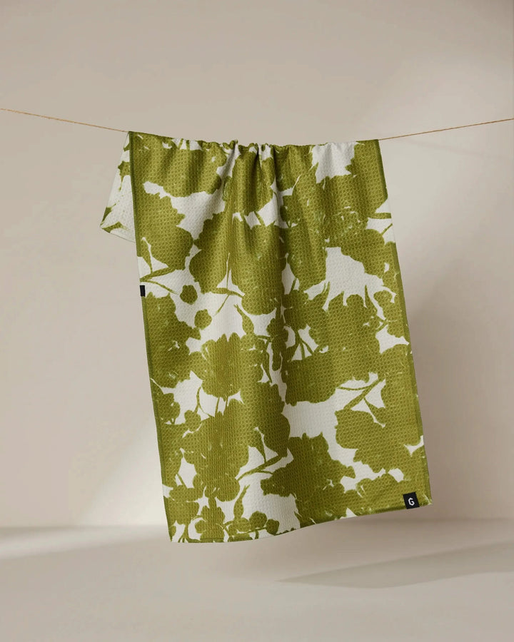 The Geometry Sweet Green tea towel, featuring a green and white abstract leaf pattern and double-sided design, hangs on a clothesline against a neutral background.