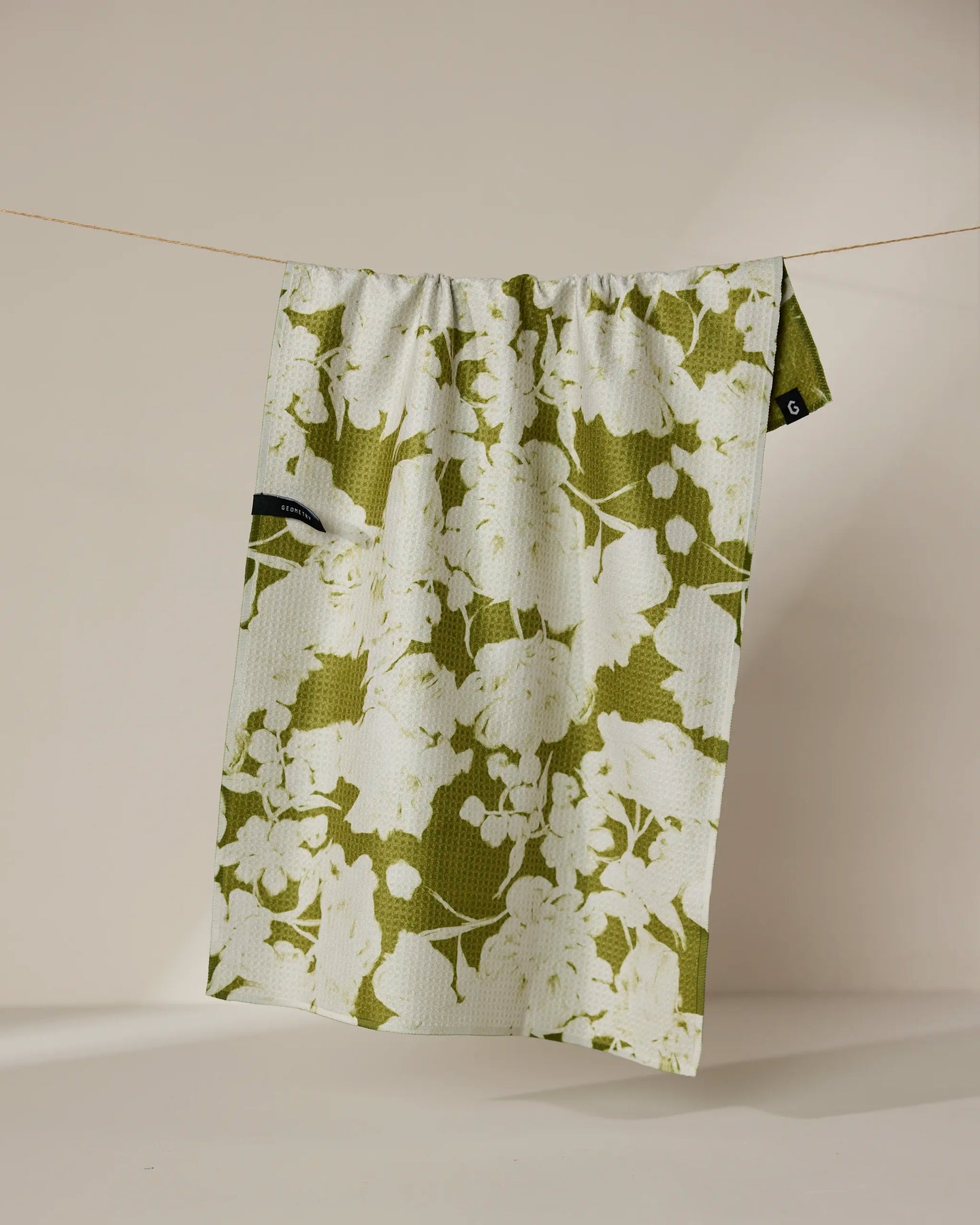 The Geometry “Sweet Green” recycled kitchen towel, featuring a green and white floral pattern and a small black tag, hangs on a thin string against a plain beige background.