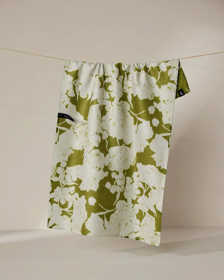 The Geometry “Sweet Green” recycled kitchen towel, featuring a green and white floral pattern and a small black tag, hangs on a thin string against a plain beige background.