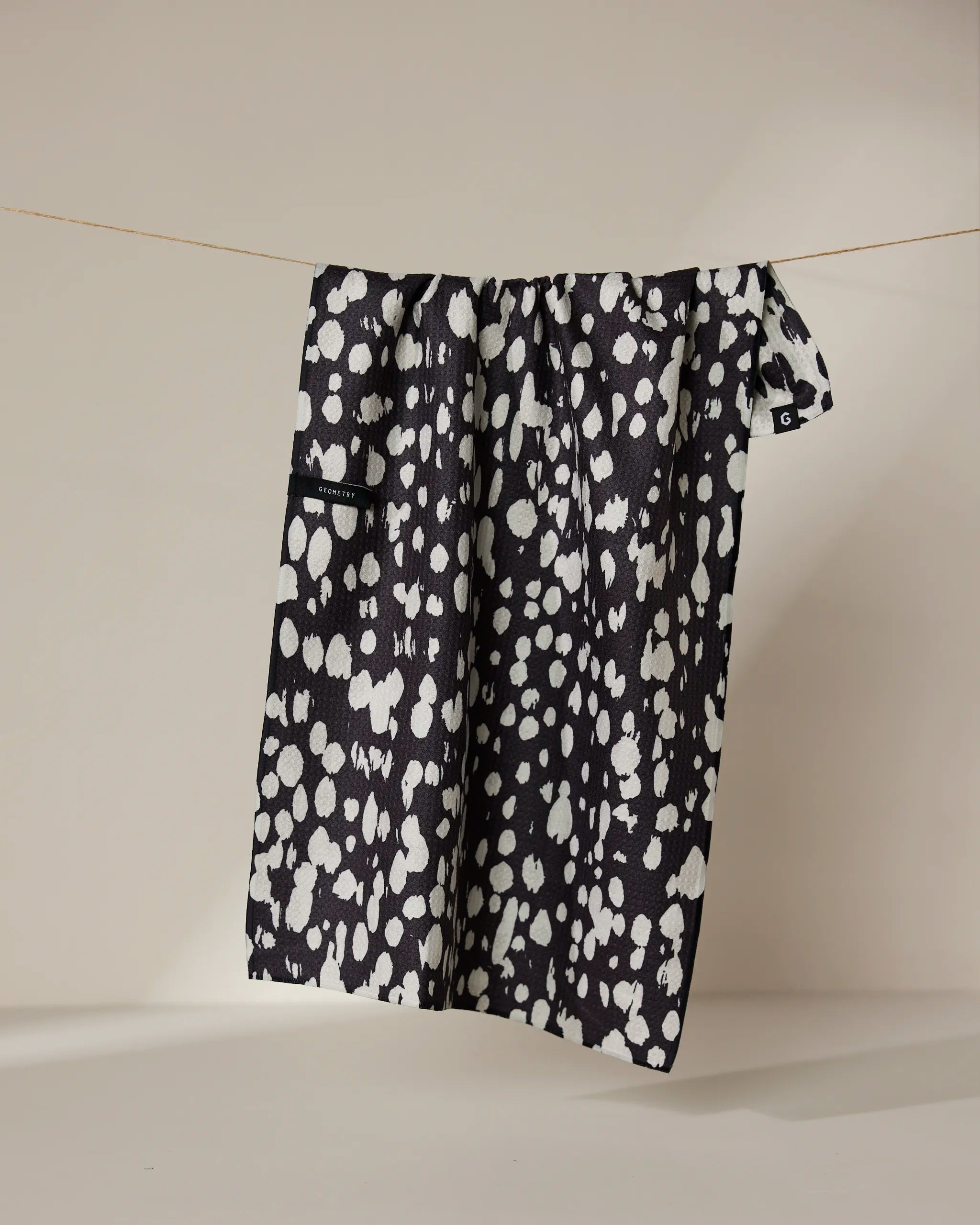 The Cass tea towel by Geometry features a black and white, double-sided irregular spotted pattern and hangs from a thin string against a neutral background.