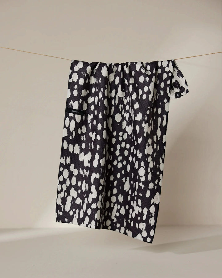 The Cass tea towel by Geometry features a black and white, double-sided irregular spotted pattern and hangs from a thin string against a neutral background.