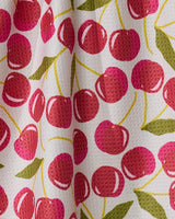 A close-up of Geometry’s Cherry Chic tea towel shows its quick-drying white textured fabric with a repeating pattern of red cherries with yellow stems and green leaves, softly draped to create gentle folds.