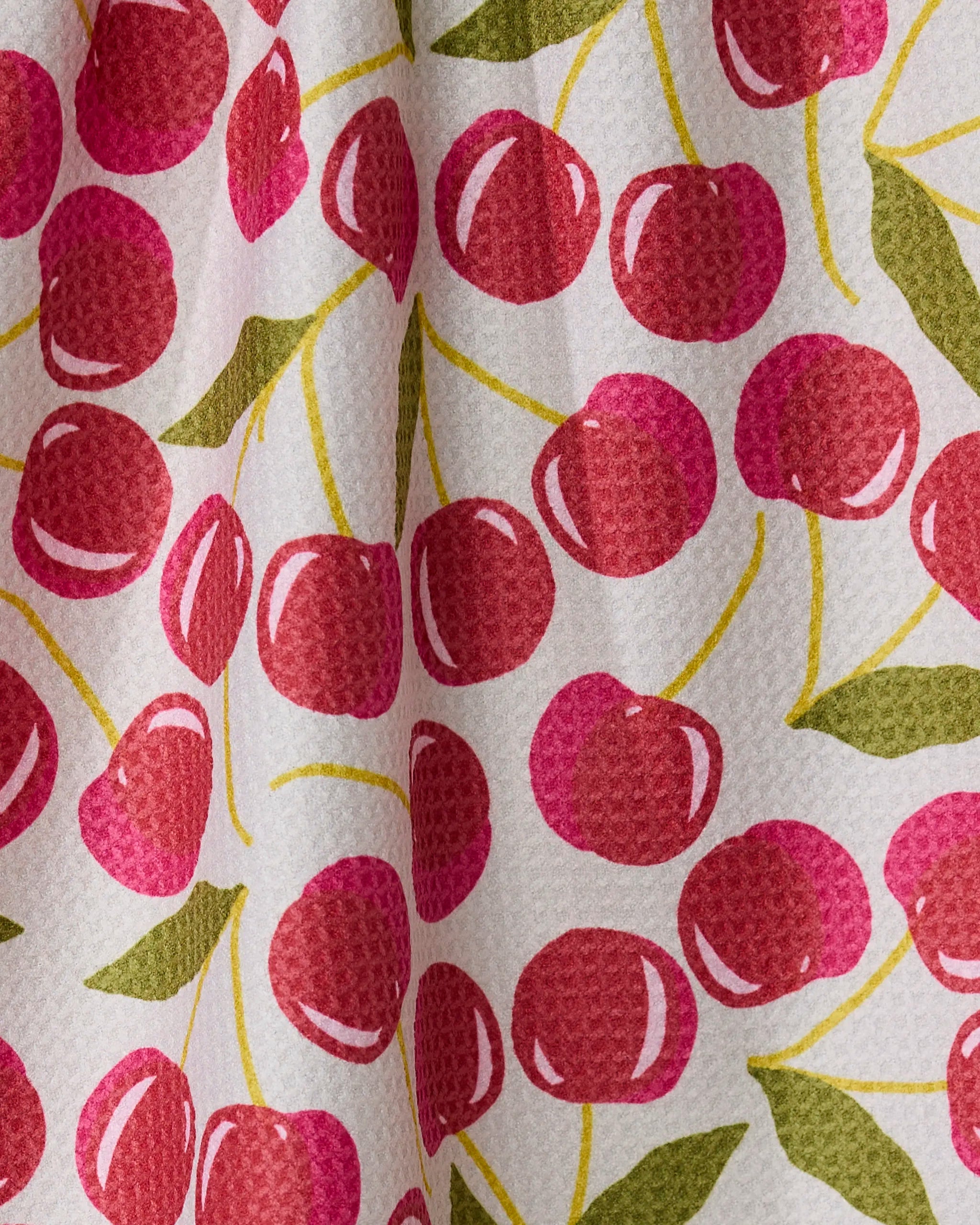 A close-up of Geometry’s Cherry Chic tea towel shows its quick-drying white textured fabric with a repeating pattern of red cherries with yellow stems and green leaves, softly draped to create gentle folds.